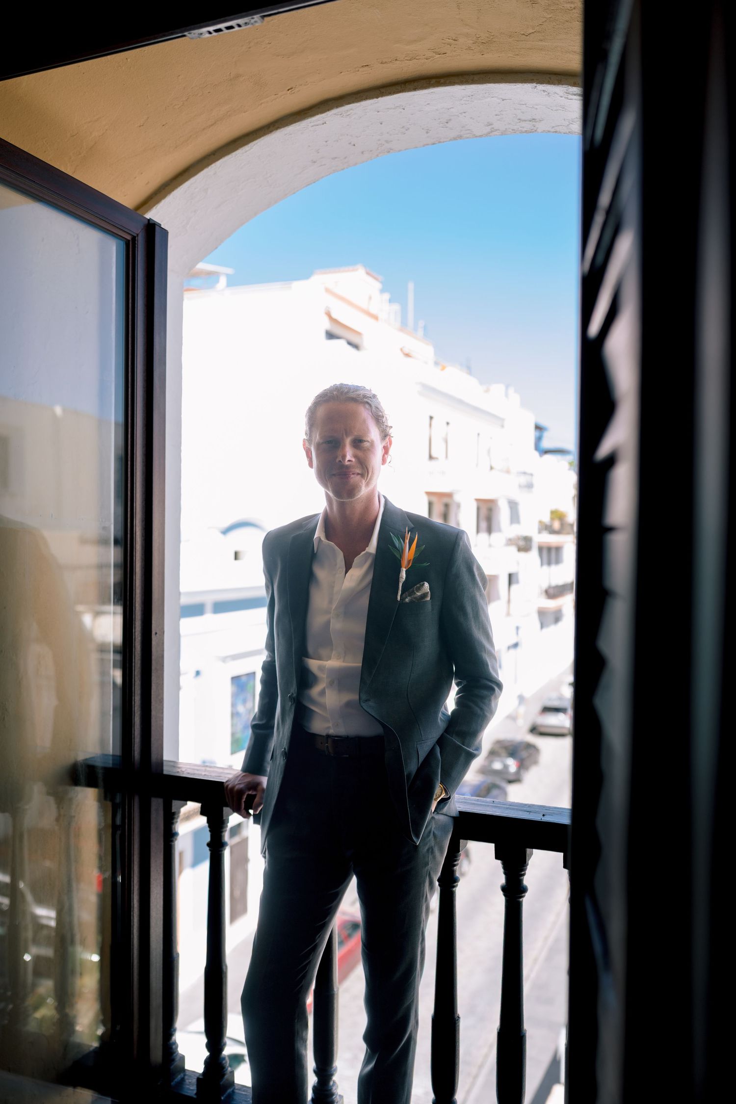 Groom in dark suit with boutonniere stands on sunlit balcony framed by arched doorway in Mediterranean town.