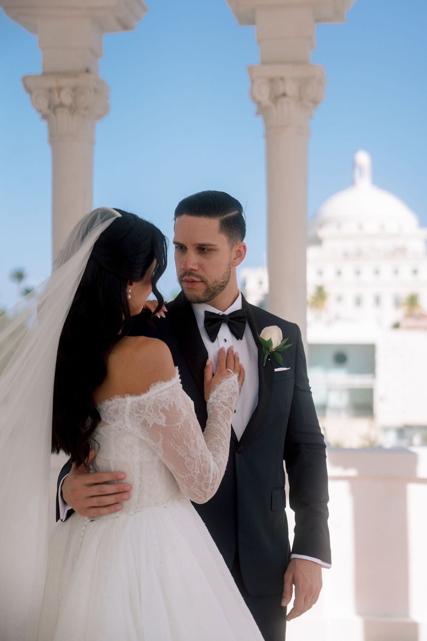 Bride in lace off-shoulder gown and groom in black tuxedo share romantic moment on elegant white columned balcony.