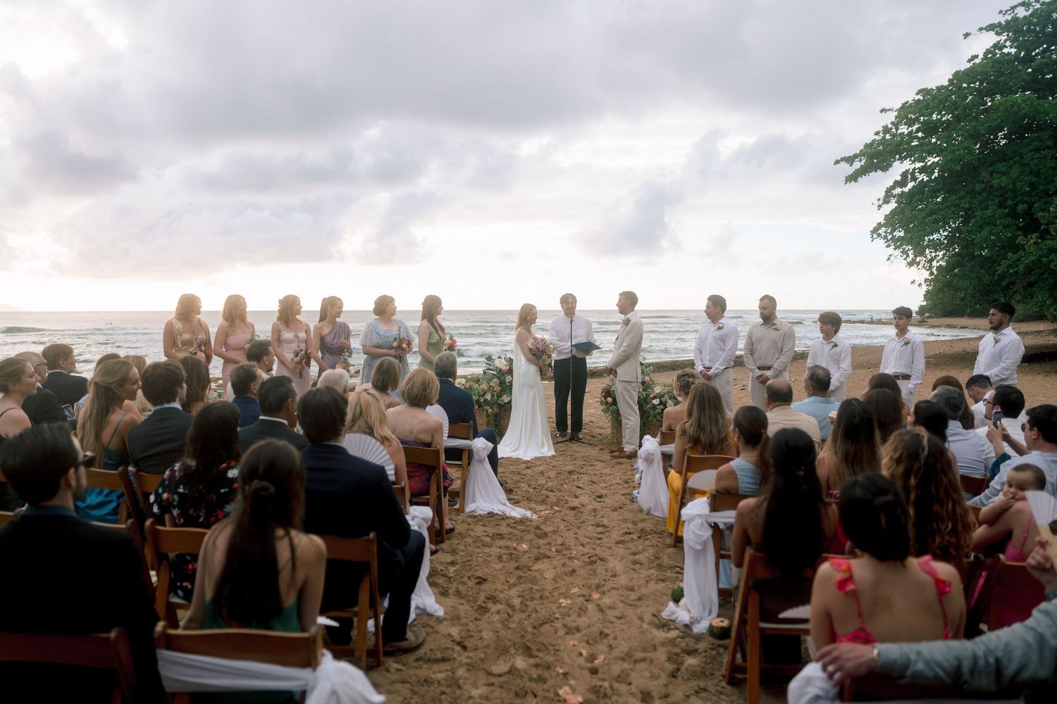 Outdoor beach wedding ceremony with guests seated on wooden chairs as couple exchanges vows by the ocean at sunset.