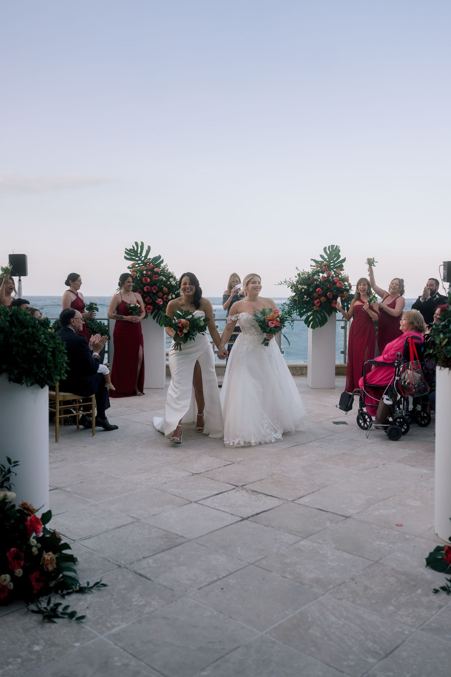 Two brides in white gowns walk down the aisle at an outdoor oceanfront wedding ceremony with red floral arrangements.