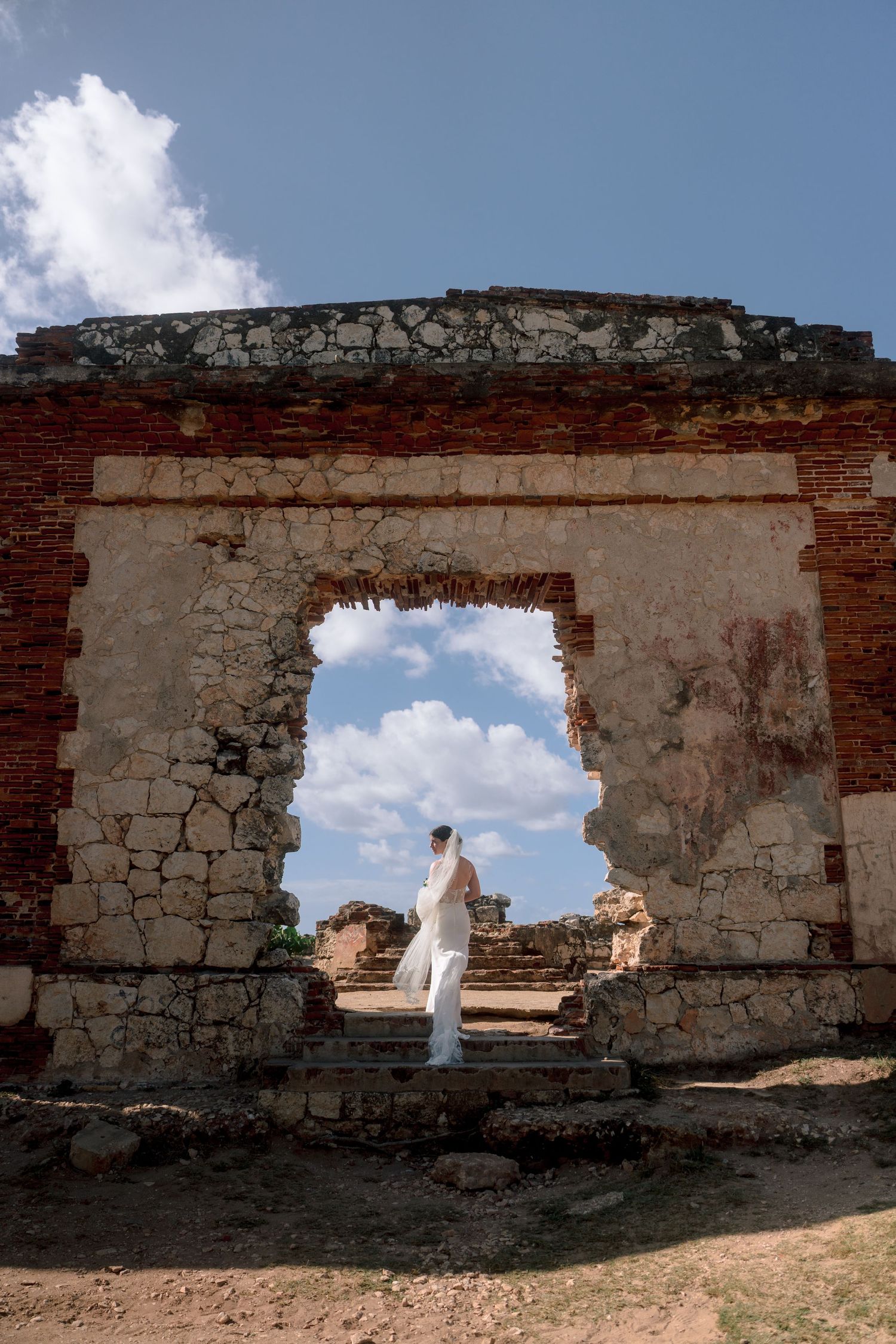 Bride in white gown stands in ancient stone archway ruins under blue sky.
