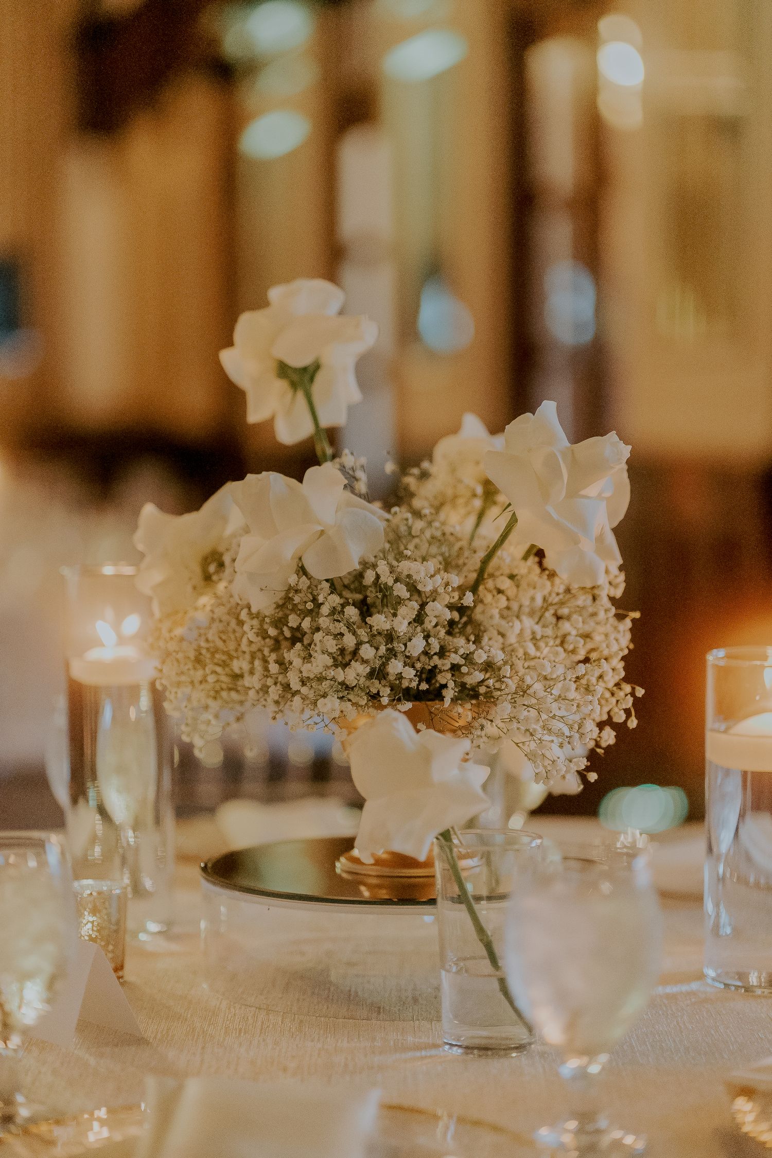 Elegant white wedding centerpiece with baby's breath and orchids on gold mirror surrounded by candles.