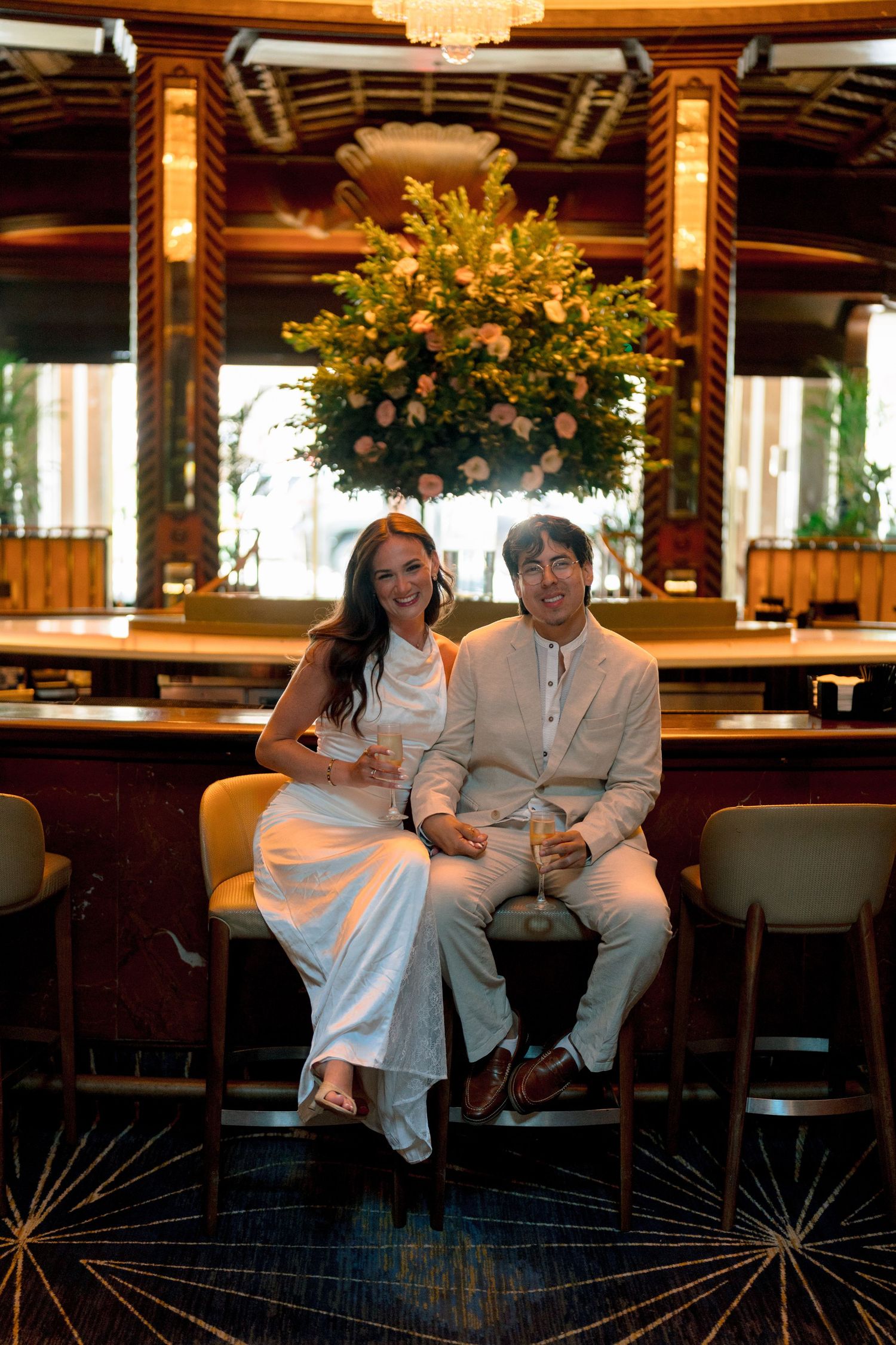 Couple in elegant attire sitting at a luxury hotel bar with floral arrangement backdrop.