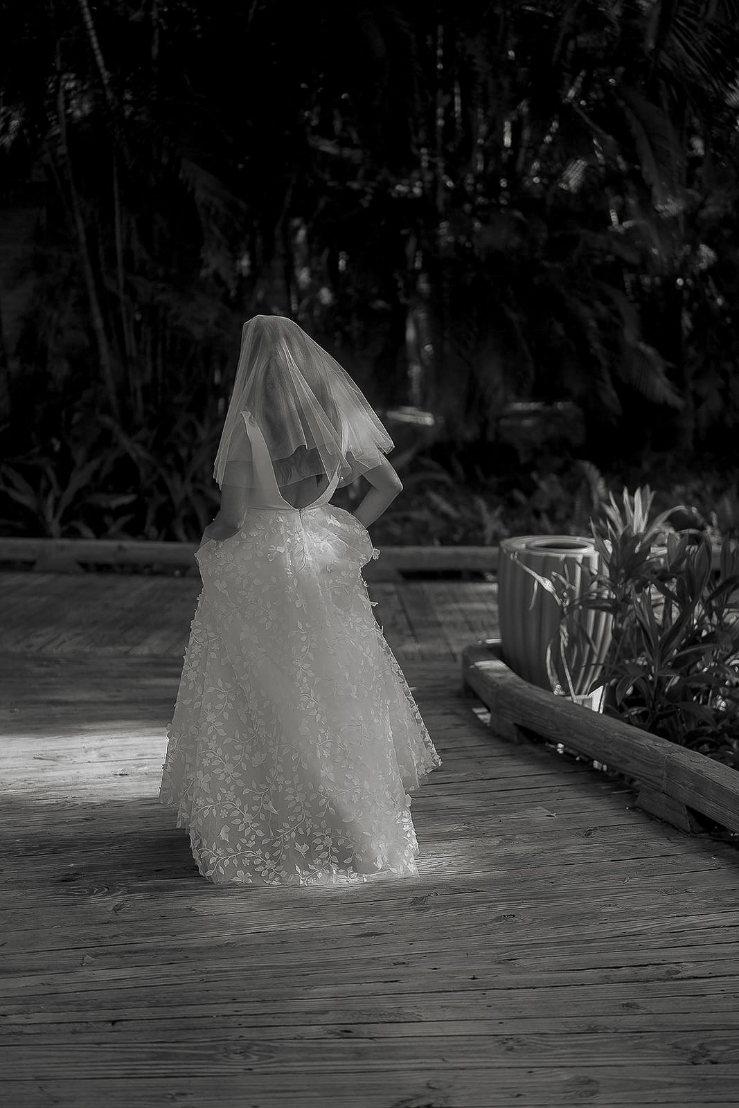 Black and white photo of a bride in a lace wedding gown and veil standing on a wooden deck surrounded by tropical plants.