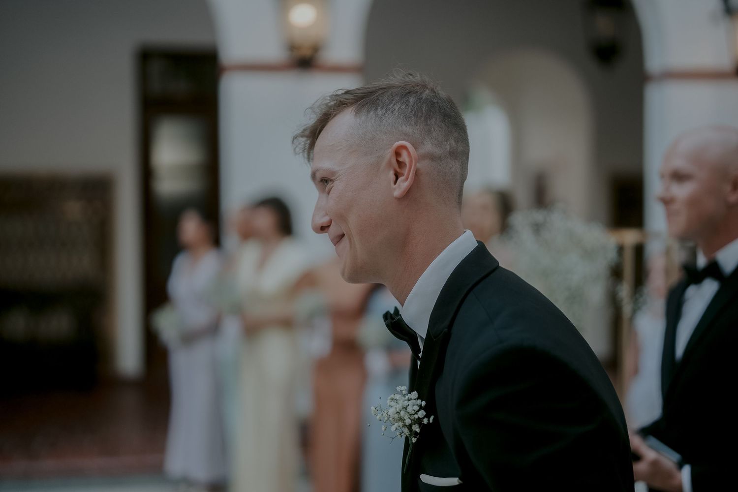 Smiling groom in black tuxedo with boutonniere at wedding ceremony, guests in elegant attire visible in background.