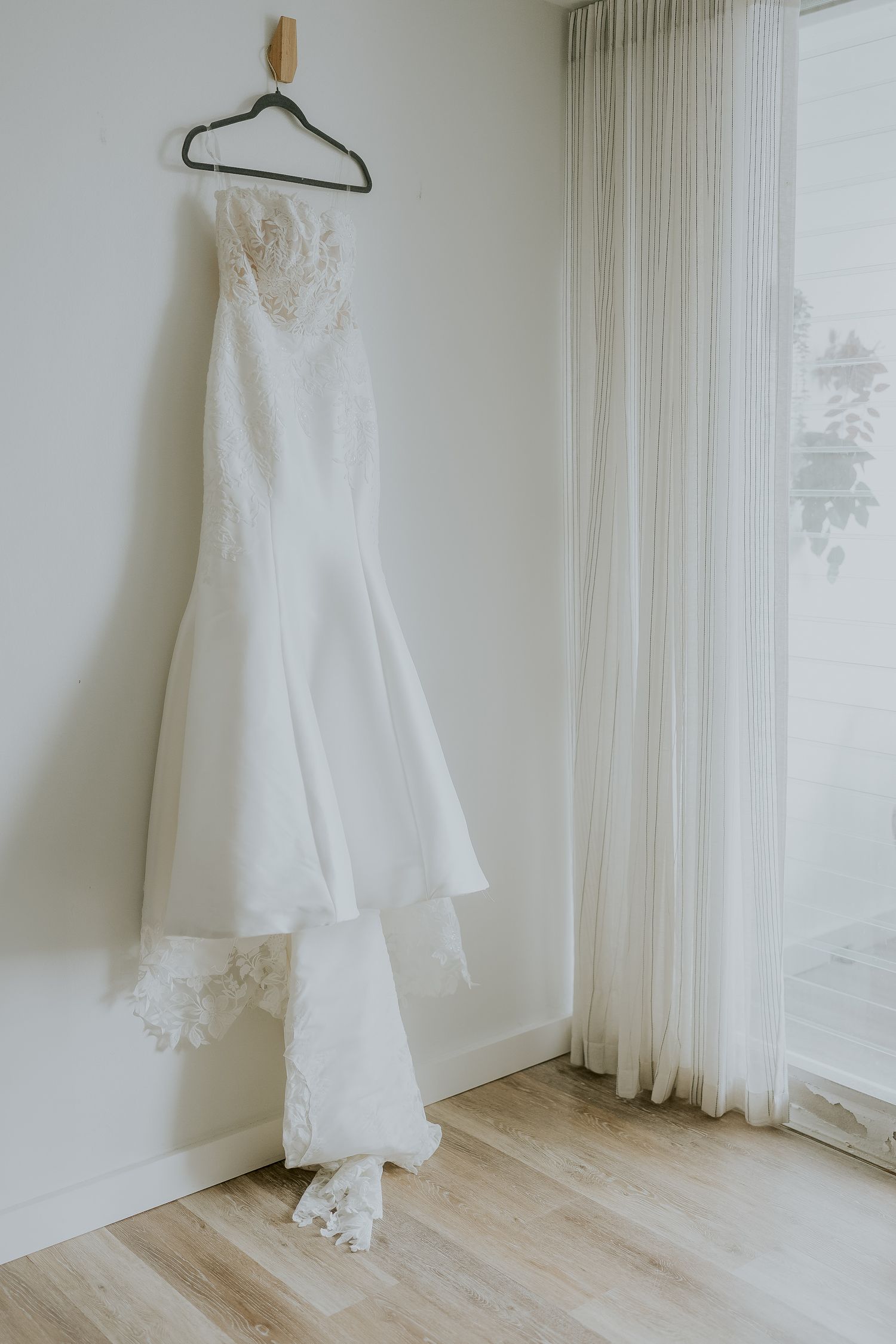 Elegant white lace wedding dress hanging on a black hanger against a bright white wall near a window.
