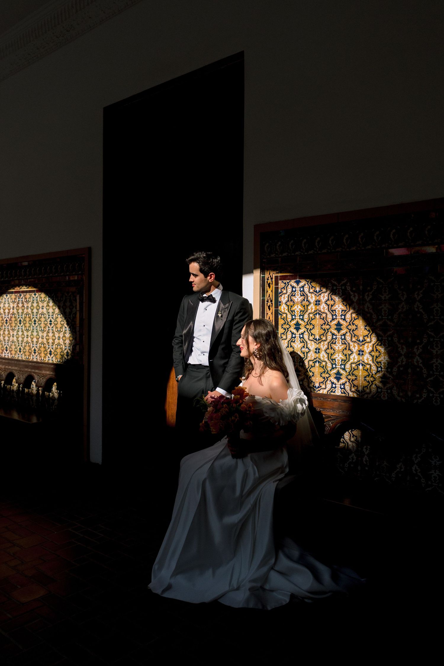Bride and groom pose dramatically in a dark ornate room with colorful tilework, bride seated in wedding gown, groom in tuxedo.