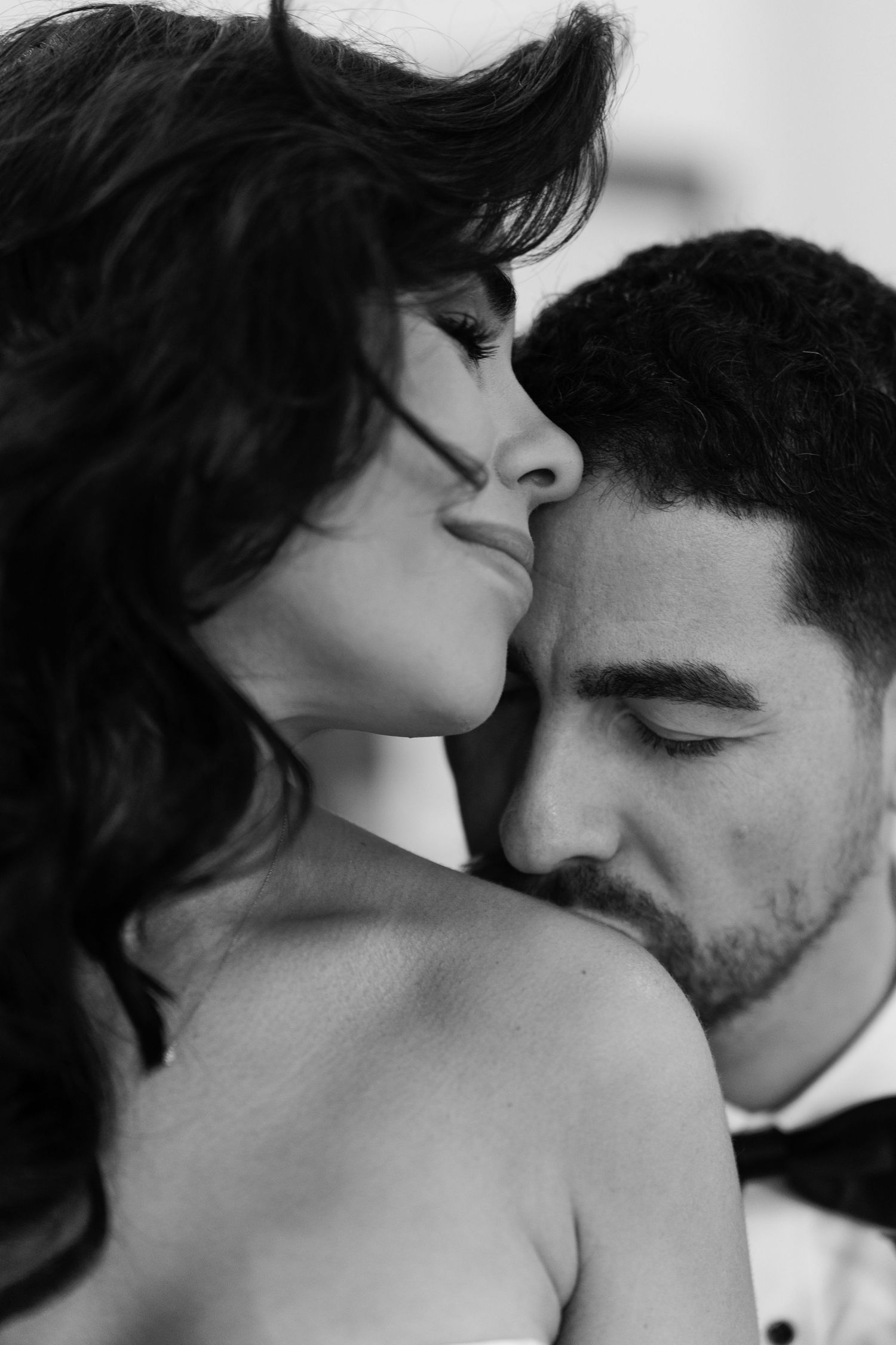 Black and white romantic photo of a couple, man in tuxedo gently kissing woman's shoulder in intimate moment.