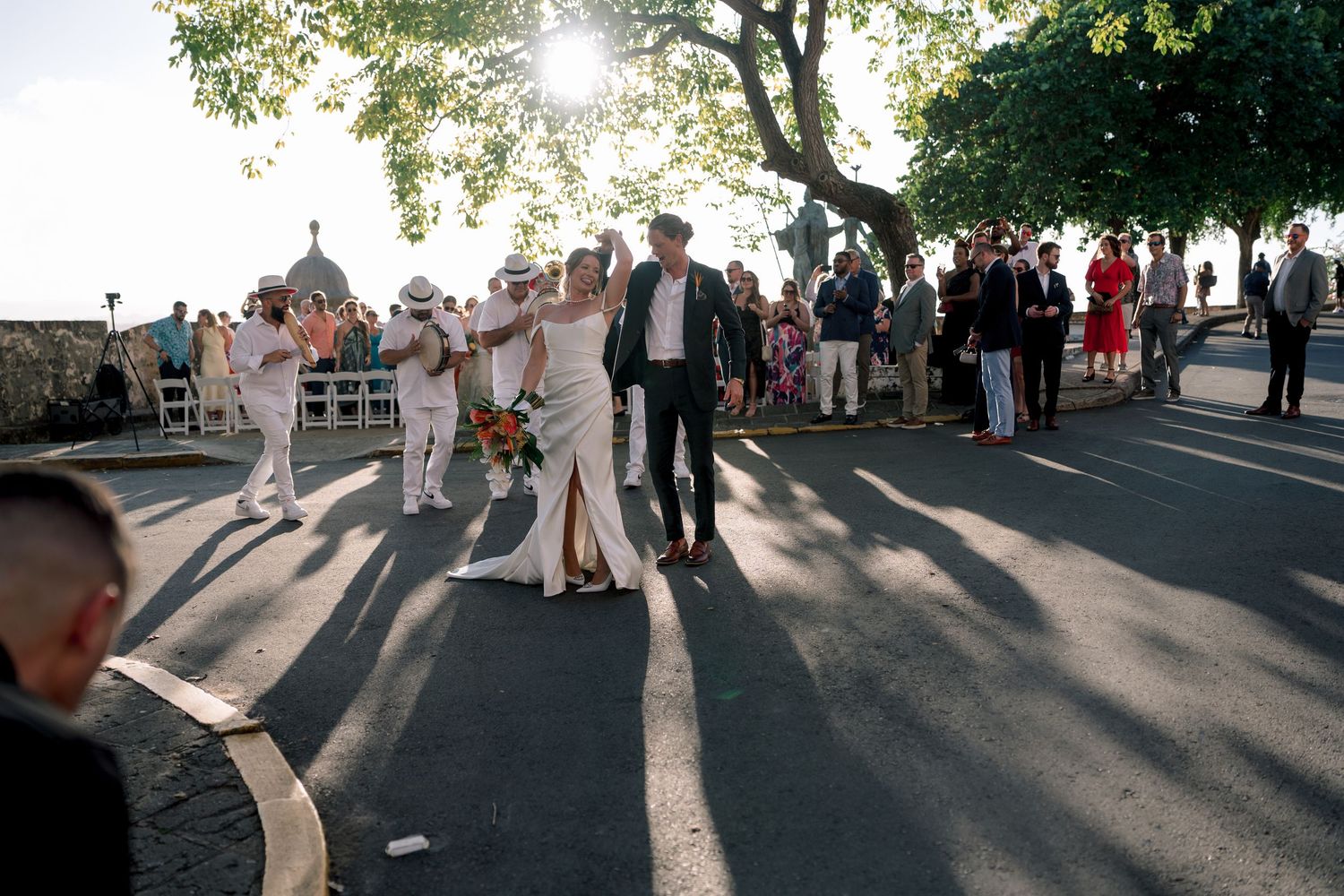 Wedding party celebrating outdoors on a sunny street, bride in white dress surrounded by guests and trees.