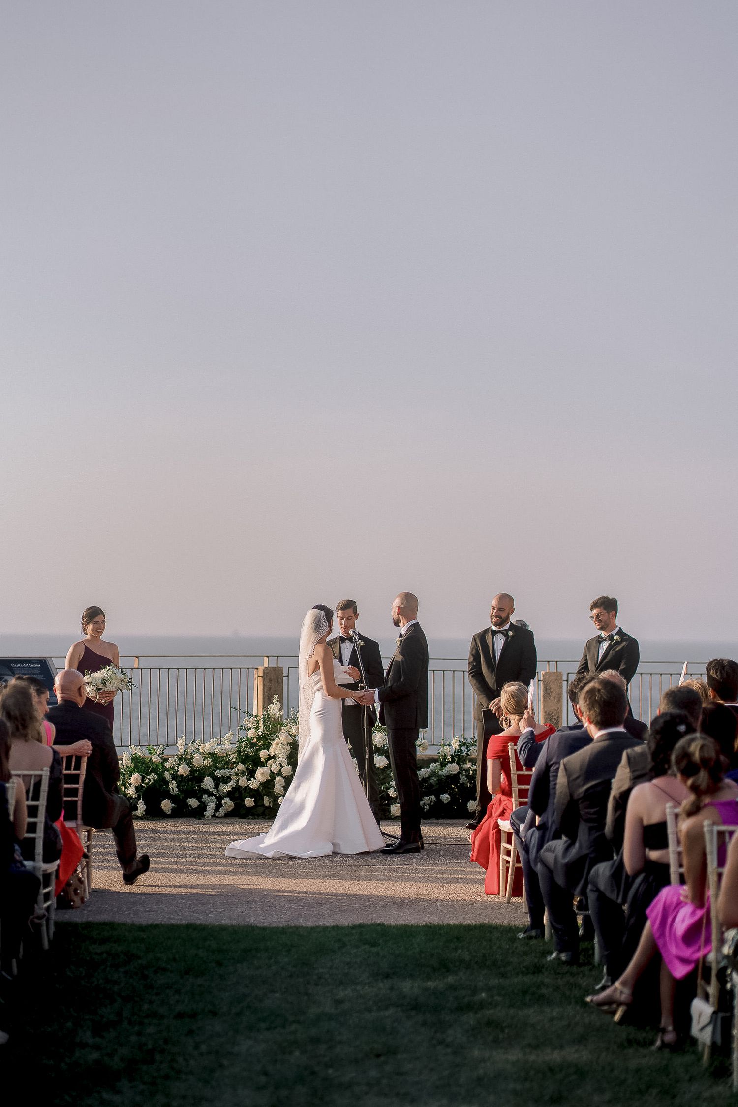 Outdoor wedding ceremony overlooking the ocean with guests seated along a white floral aisle.