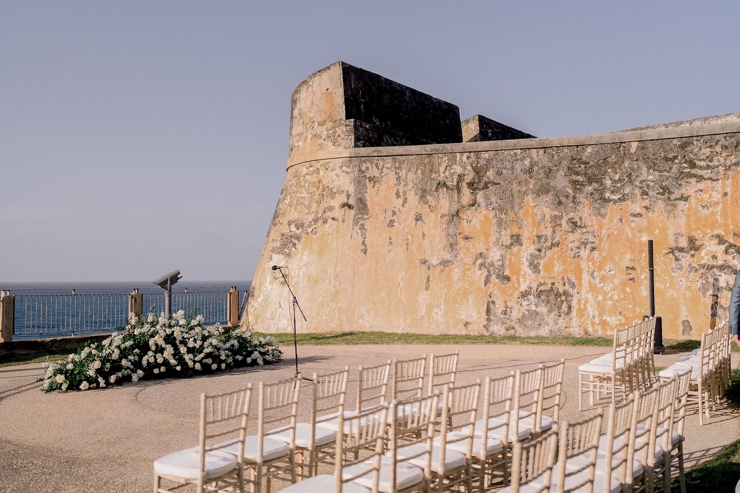 Elegant outdoor wedding ceremony setup with gold chairs beside an ancient stone fortress wall overlooking the sea.