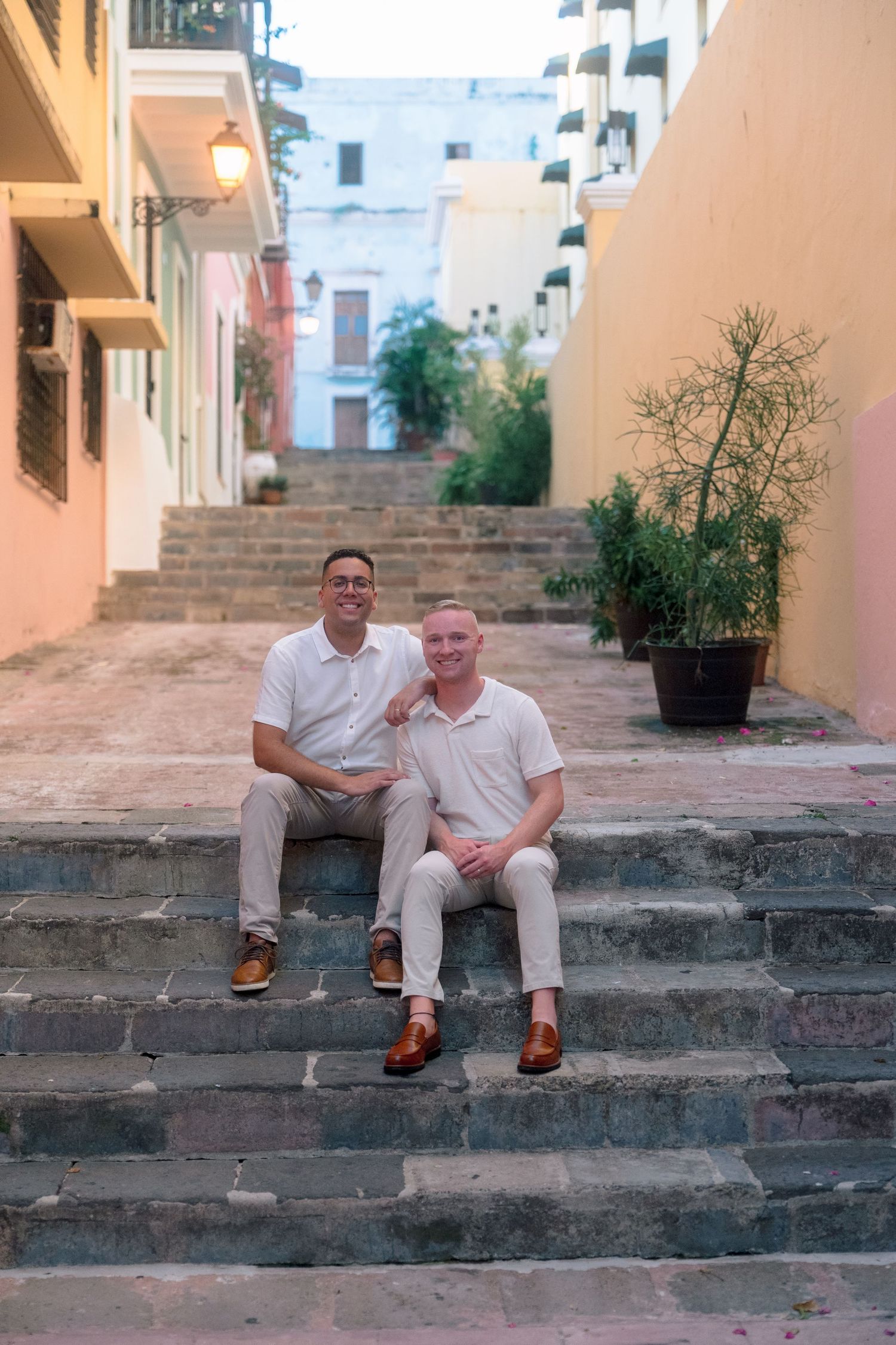 Two men in matching white shirts and khakis sit smiling on stone steps in a colorful old town alley.