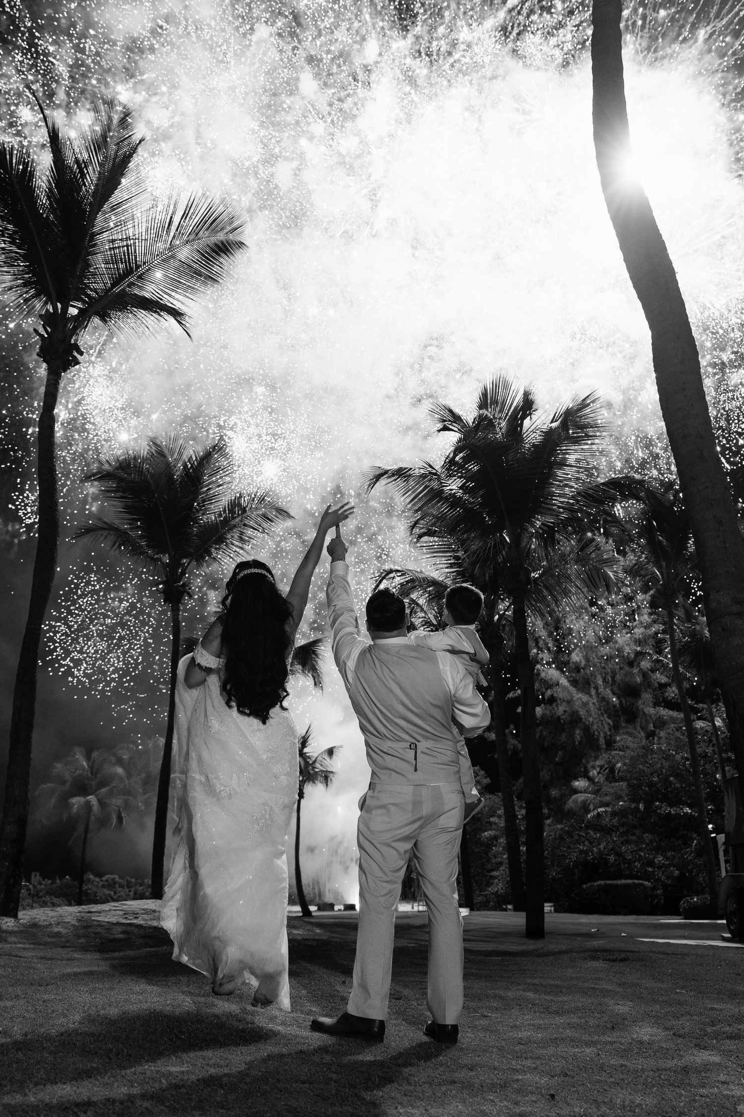 Bride and groom raise arms in celebration before spectacular fireworks display among palm trees.