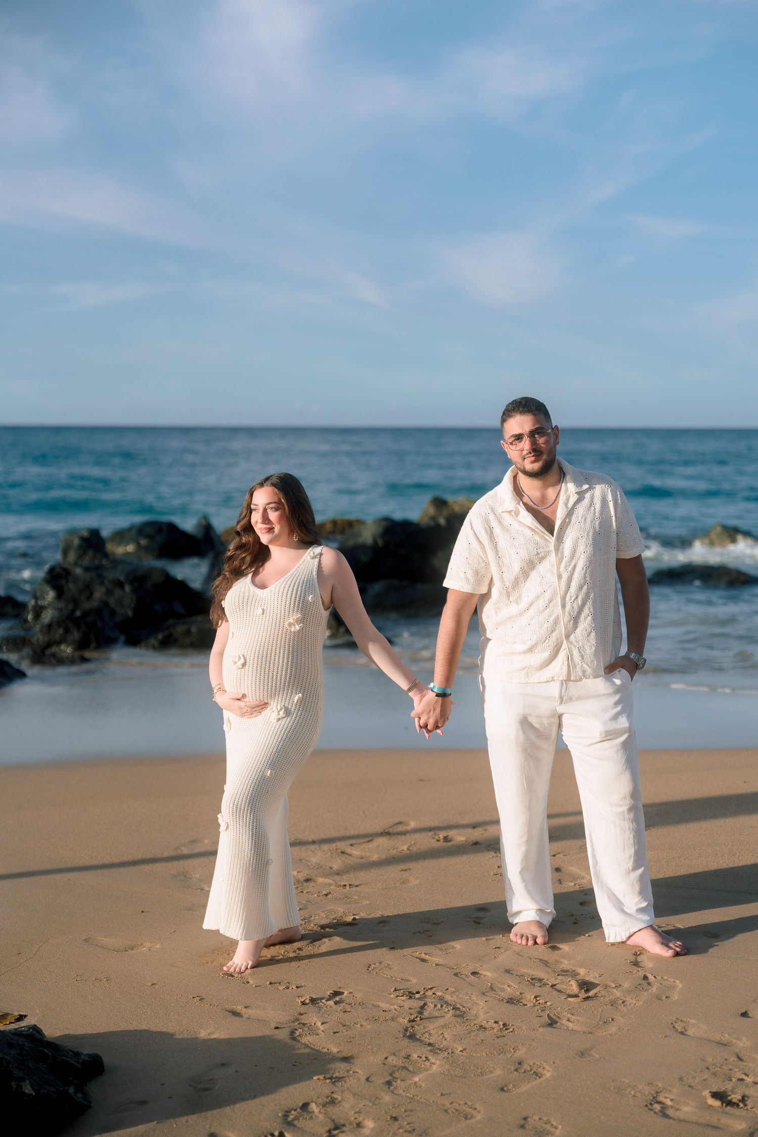 Pregnant couple holding hands on sandy beach wearing white outfits with ocean waves and rocks in background.