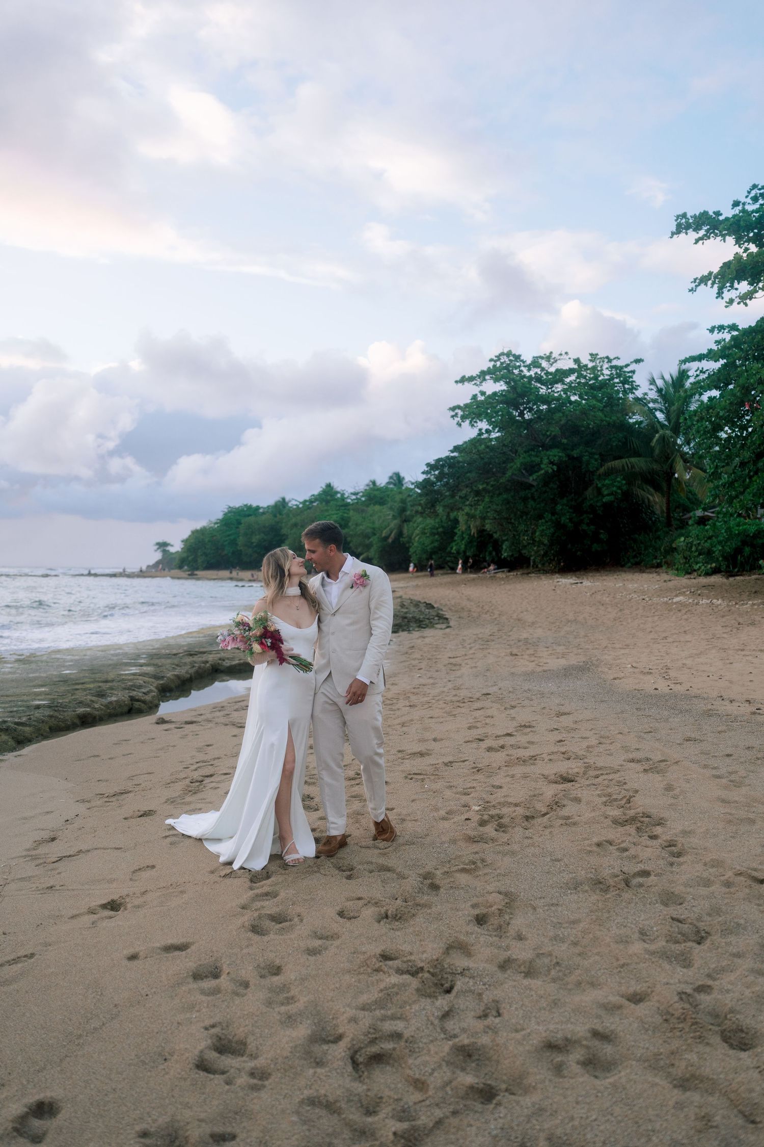Bride and groom sharing a kiss on sandy beach at dusk with lush tropical trees behind them.
