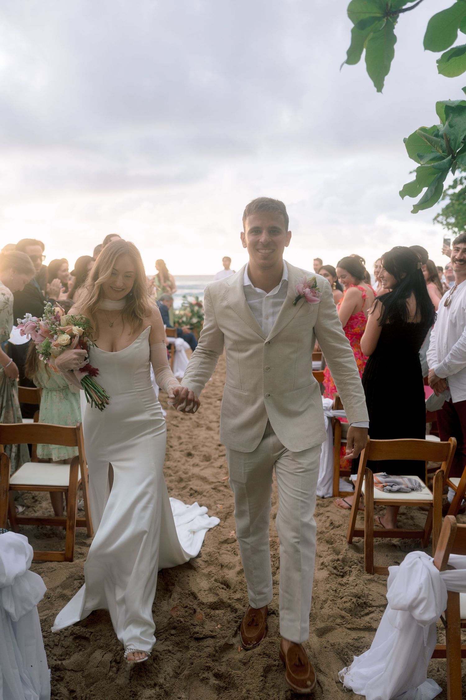 Newlyweds walk down sandy beach aisle hand in hand after sunset wedding ceremony with guests applauding.