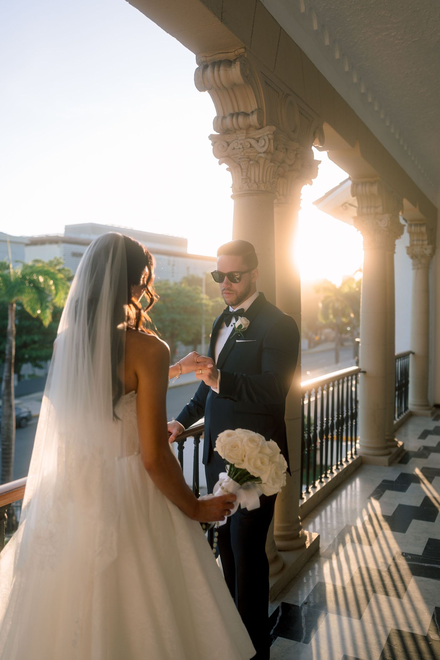 Groom in black tuxedo with sunglasses holds bride's hand on sunlit balcony, bride carries white rose bouquet.