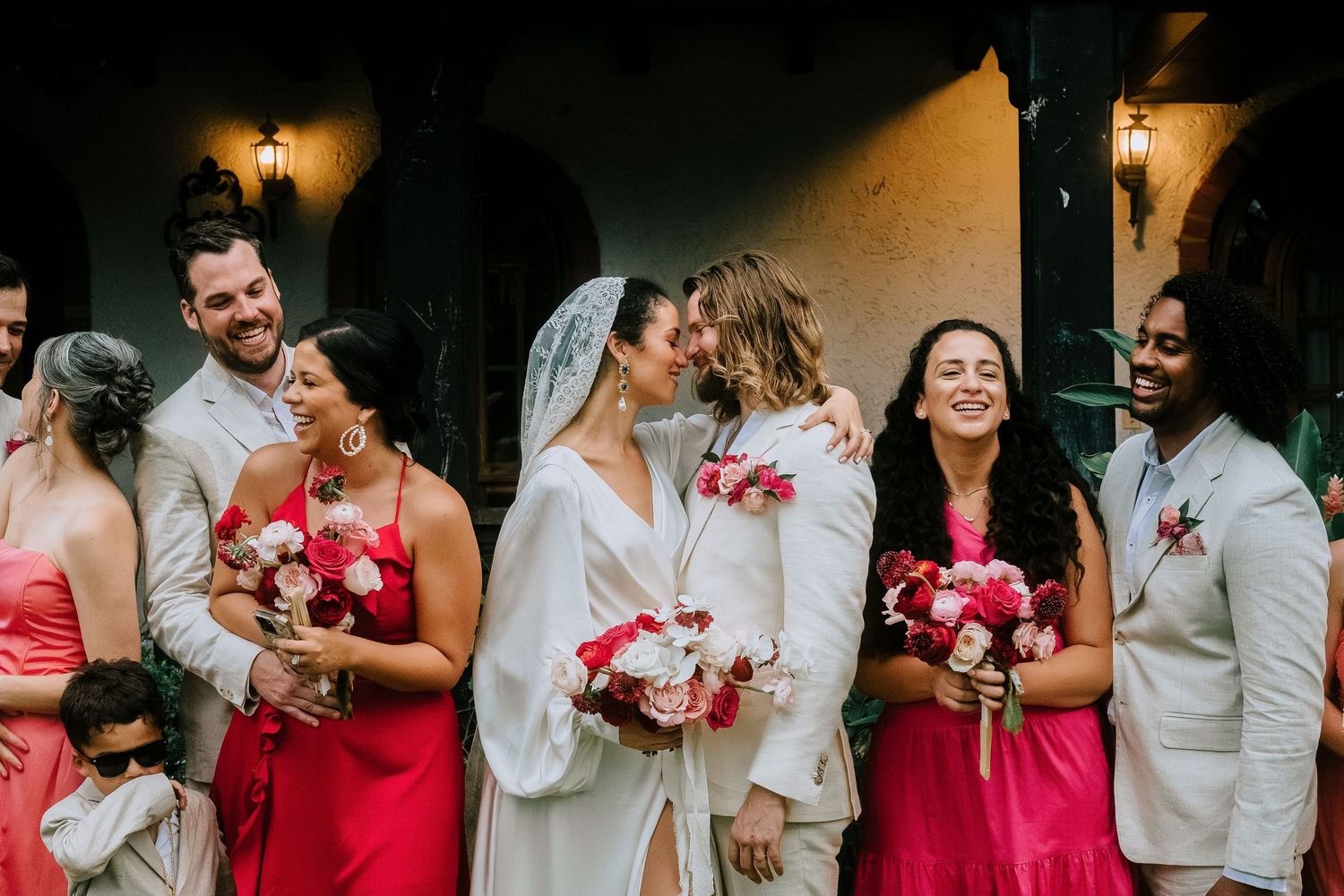 Bride and groom kiss surrounded by smiling wedding party in pink dresses outdoors.