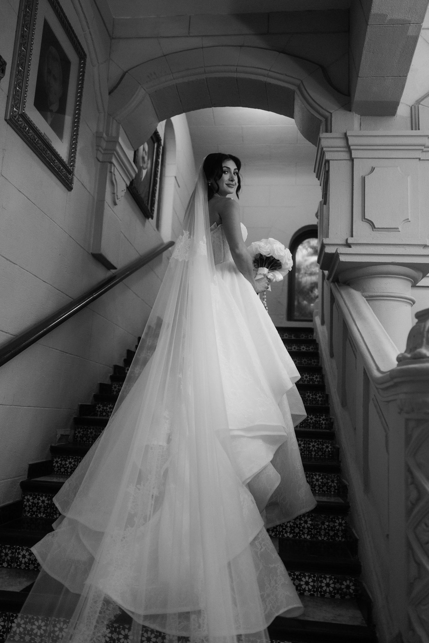 Black and white photo of bride in layered gown descending ornate staircase.