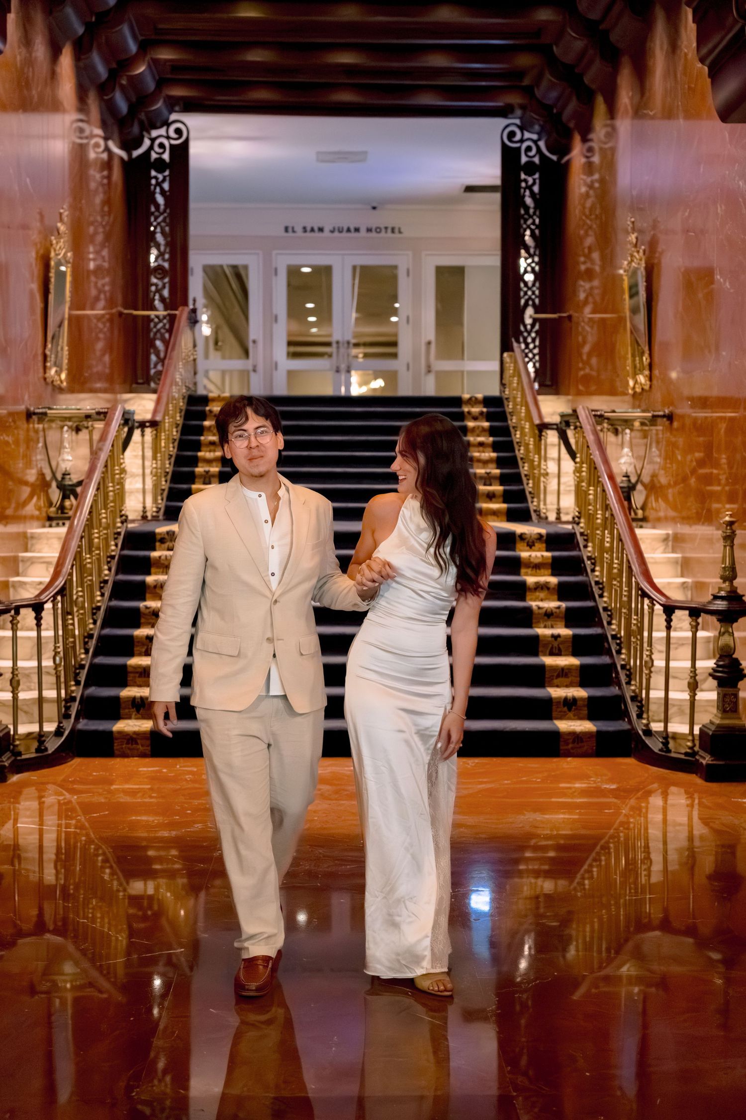 Elegant couple in white attire walking through the grand lobby of El San Juan Hotel with marble floors and ornate staircase.