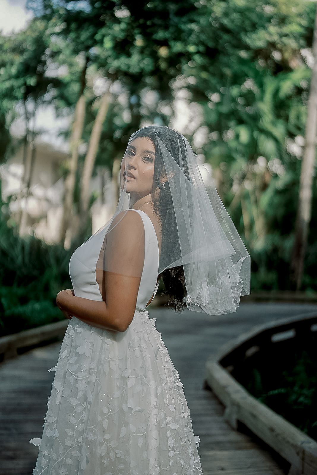 Bride in white floral gown and veil poses on wooden path surrounded by lush greenery.