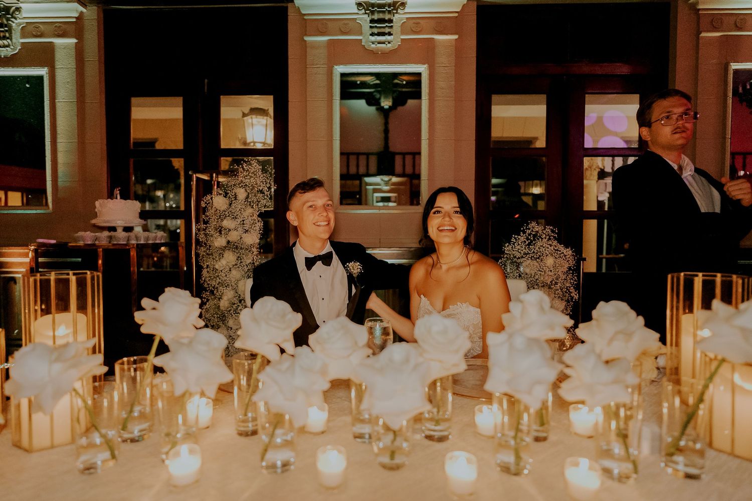 Bride and groom seated at sweetheart table with white roses and gold candle centerpieces.