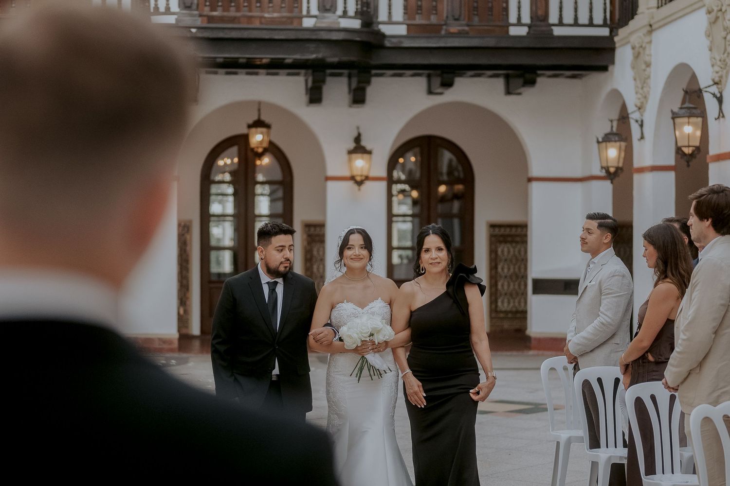 Bride walks down the aisle with family at an elegant outdoor wedding ceremony at a Spanish-style venue.