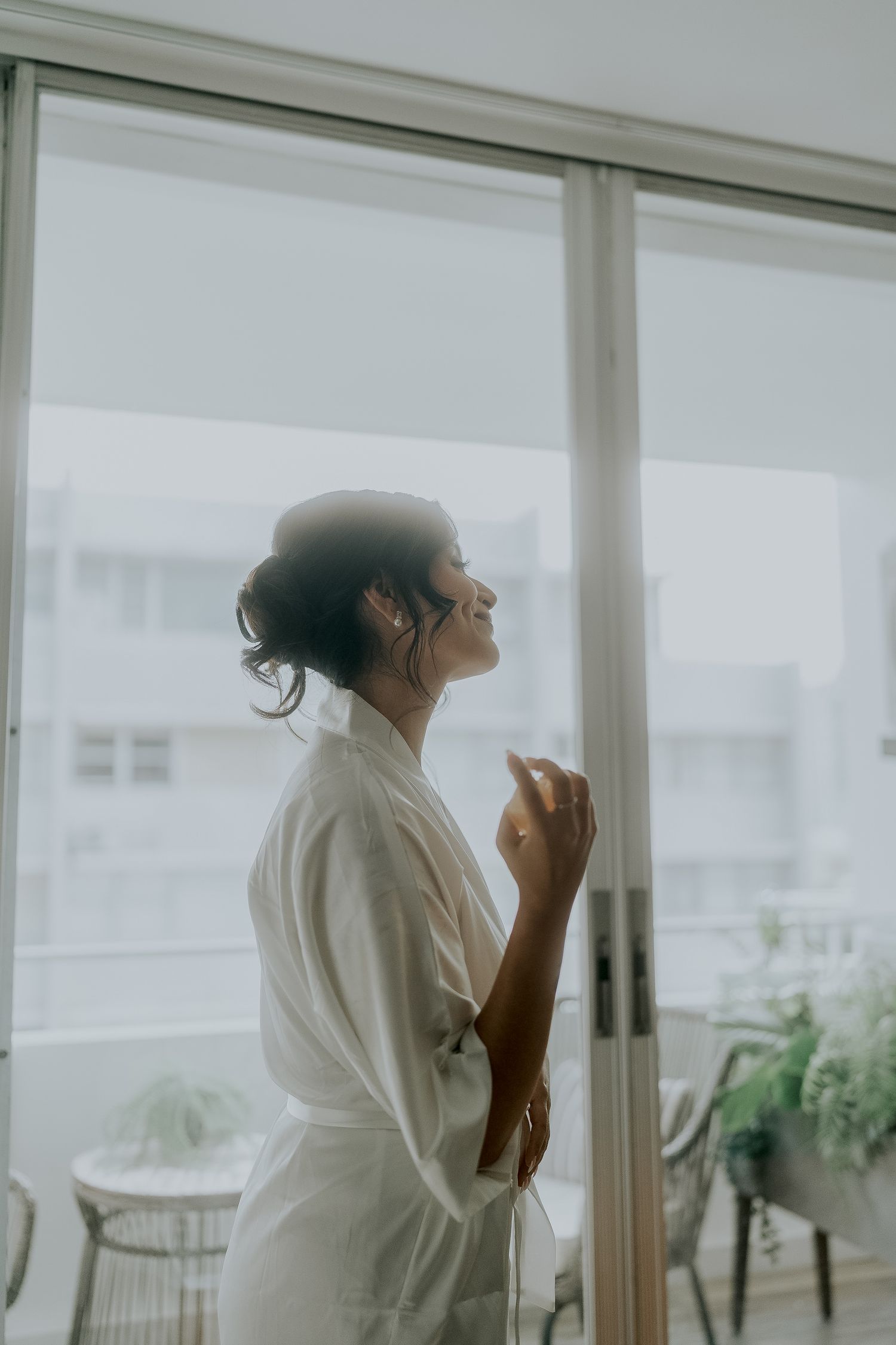 Bride in white robe smiling by large window with natural light on wedding morning.