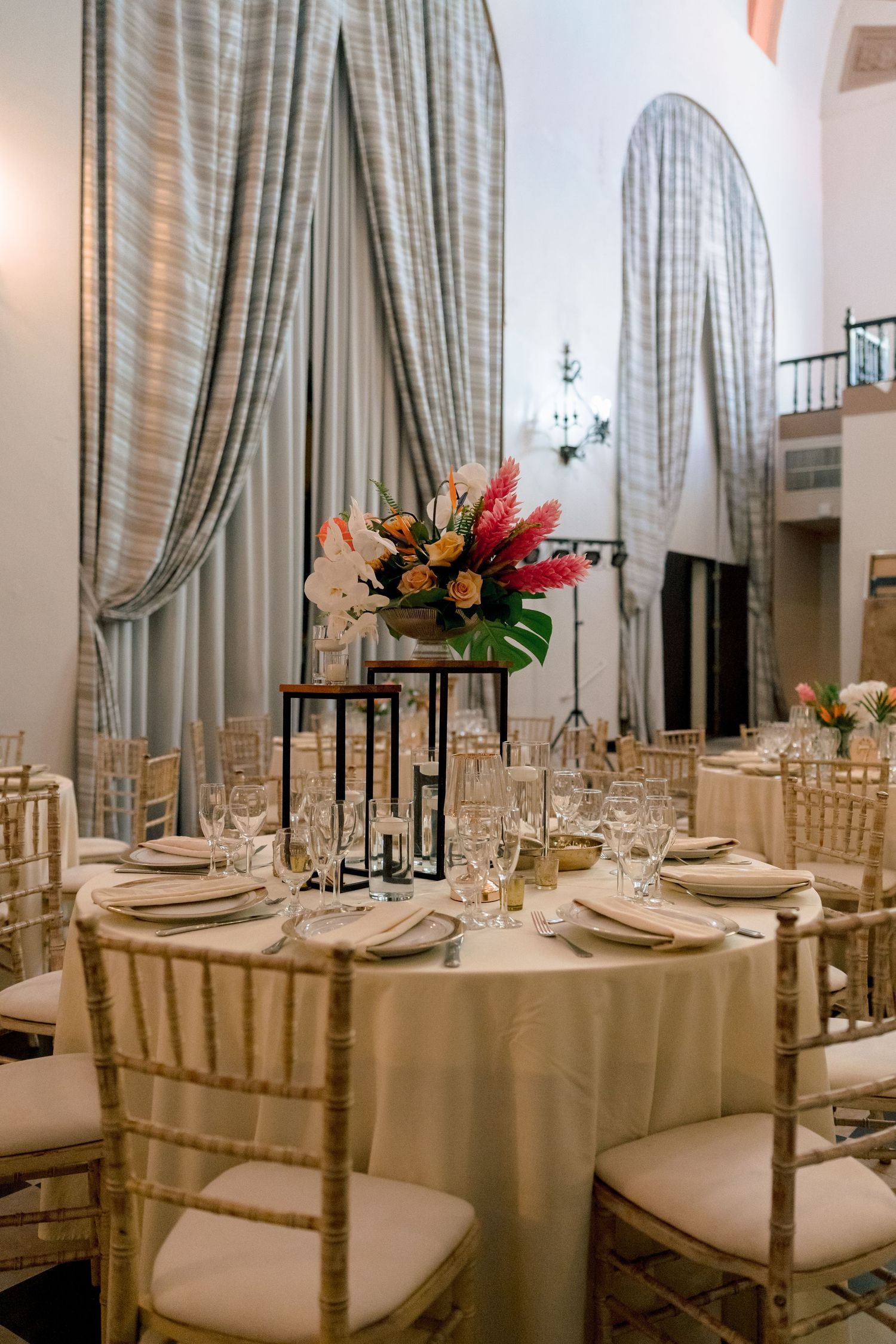 Elegant wedding reception hall with round tables, chiavari chairs, and tropical floral centerpieces in a grand venue.