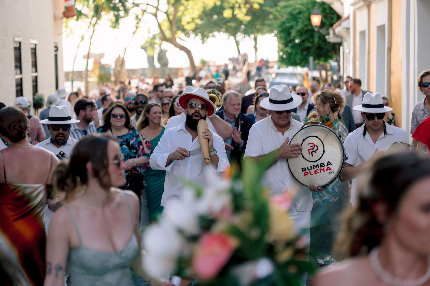 Festive wedding parade through colorful streets with guests in summer attire and live drummers performing.