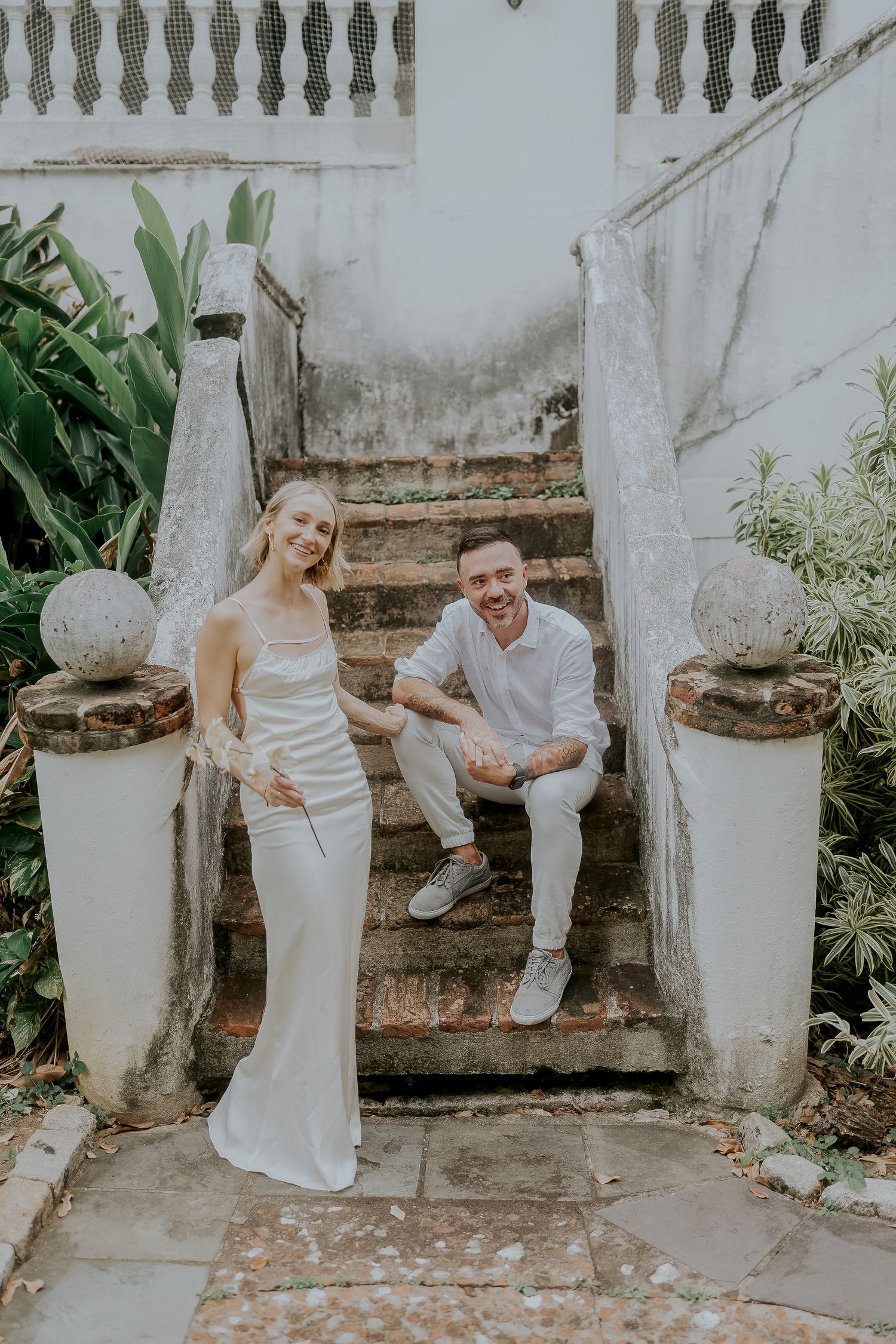 Couple in white attire smiling on rustic stone steps surrounded by lush greenery at an outdoor venue.