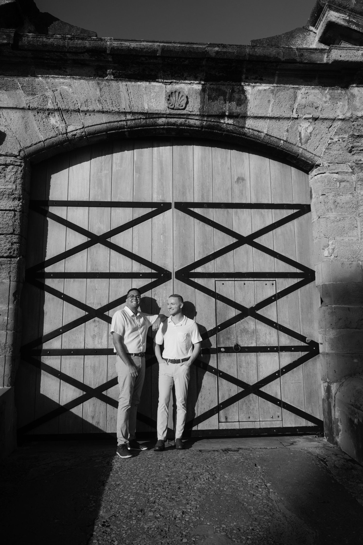 Black and white photo of two men posing in front of a large wooden arched gateway in a historic stone wall.