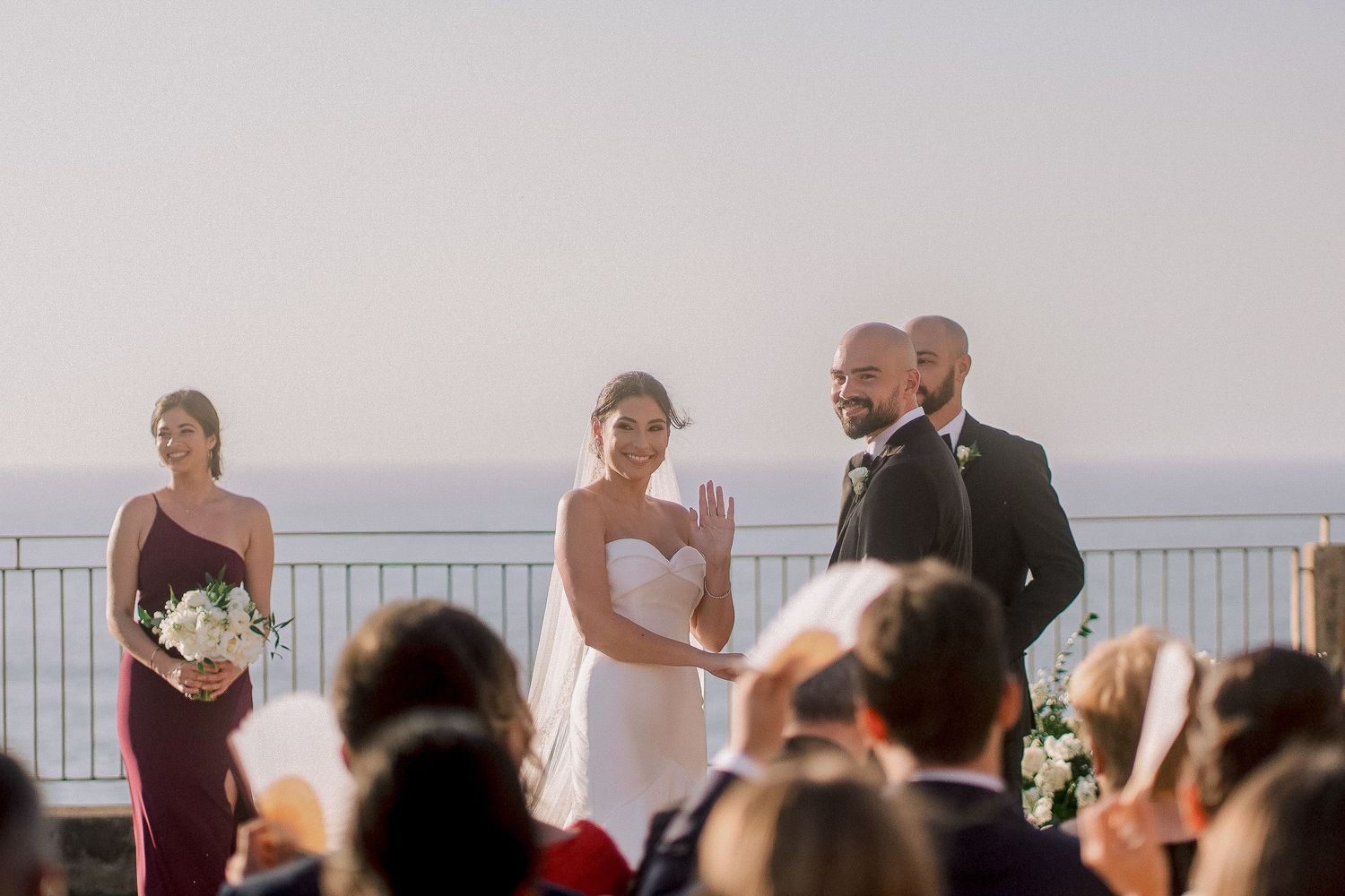 Bride and groom exchange vows at outdoor wedding ceremony with ocean view backdrop and bridesmaid in burgundy dress.