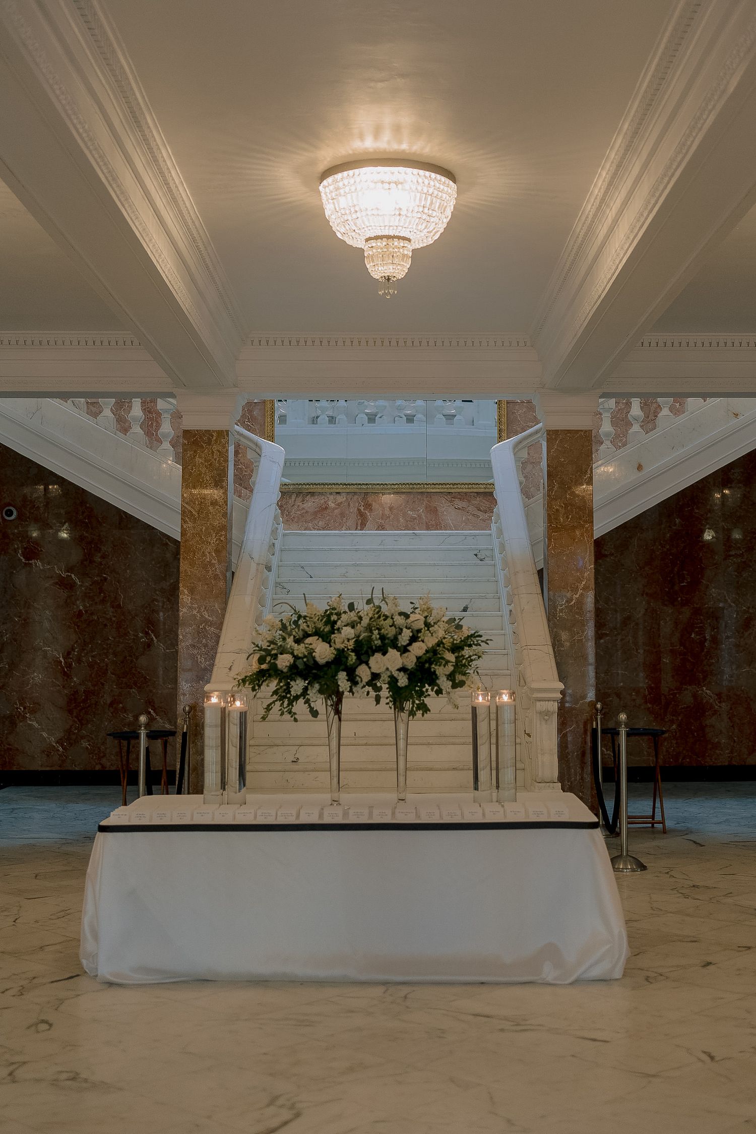 Elegant wedding reception table with white floral arrangement and candles in a marble foyer with grand staircase.
