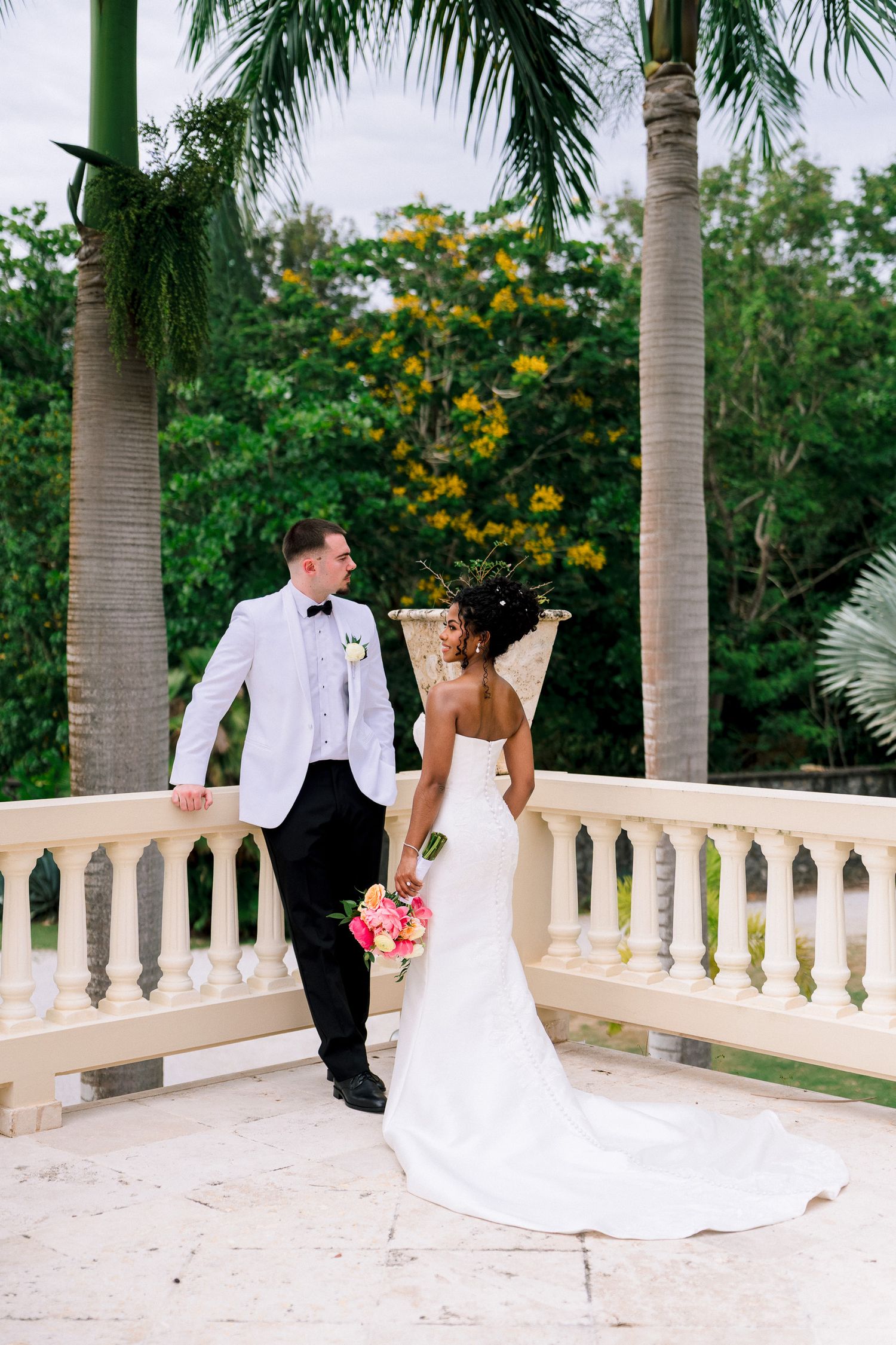 Bride and groom share a romantic moment on a balcony surrounded by tropical palm trees at their outdoor wedding.