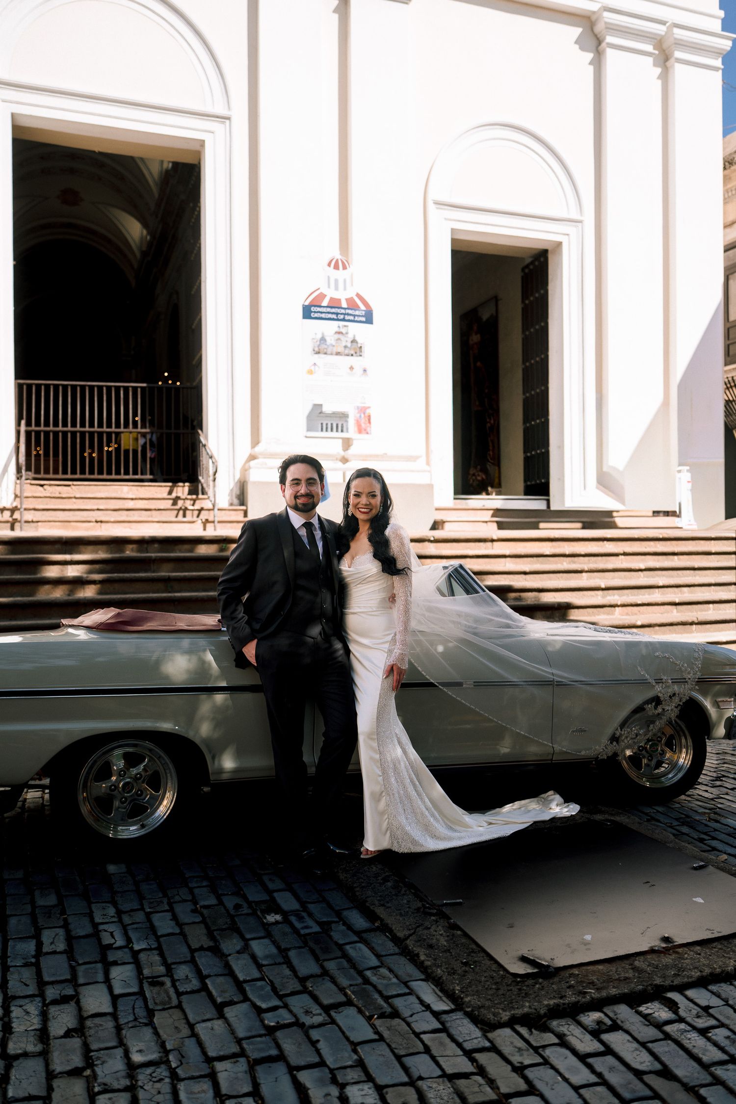 Bride and groom pose beside a vintage convertible car on cobblestone street outside a white church.