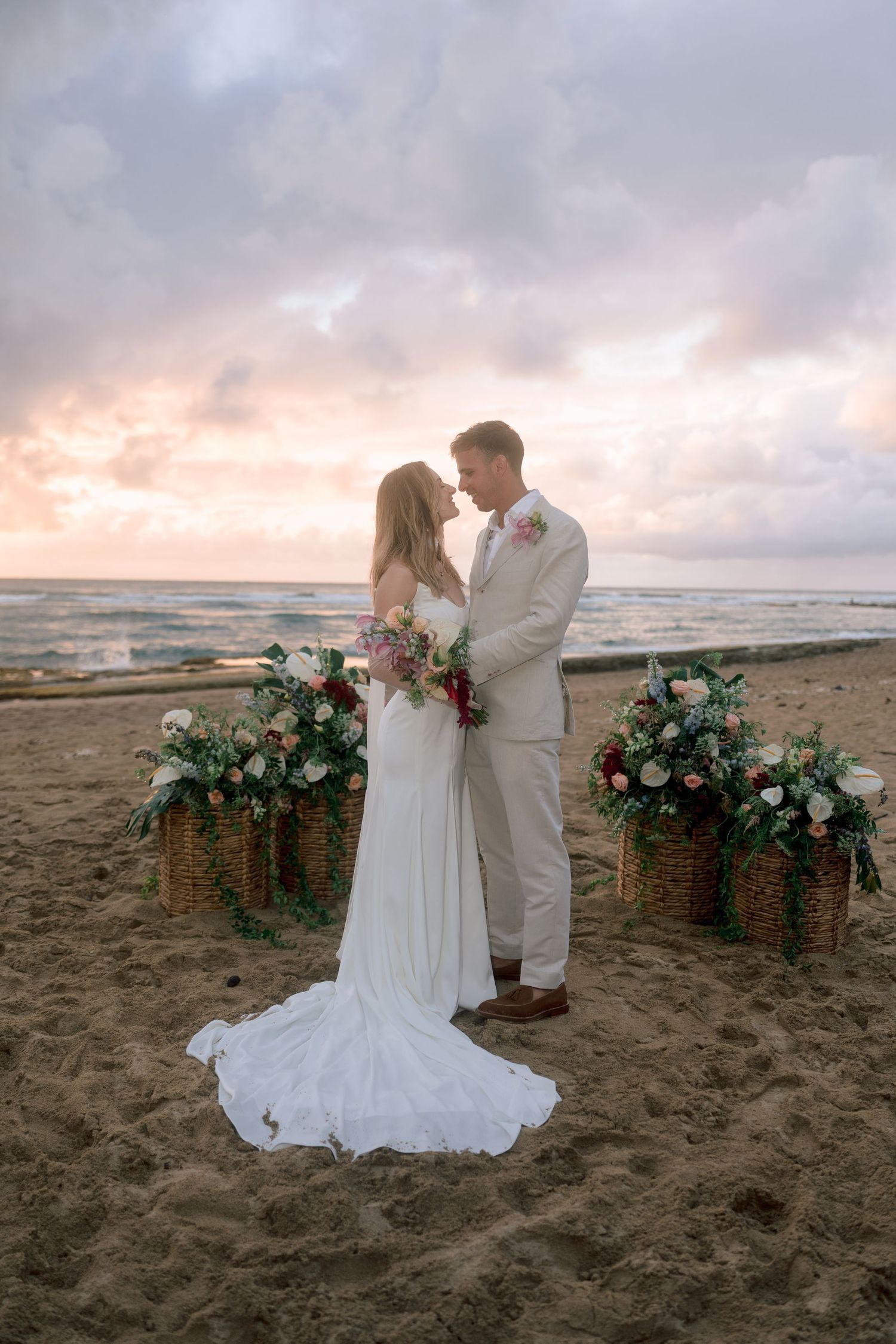 Newlyweds share intimate moment at beach wedding ceremony with colorful sunset and ocean backdrop.