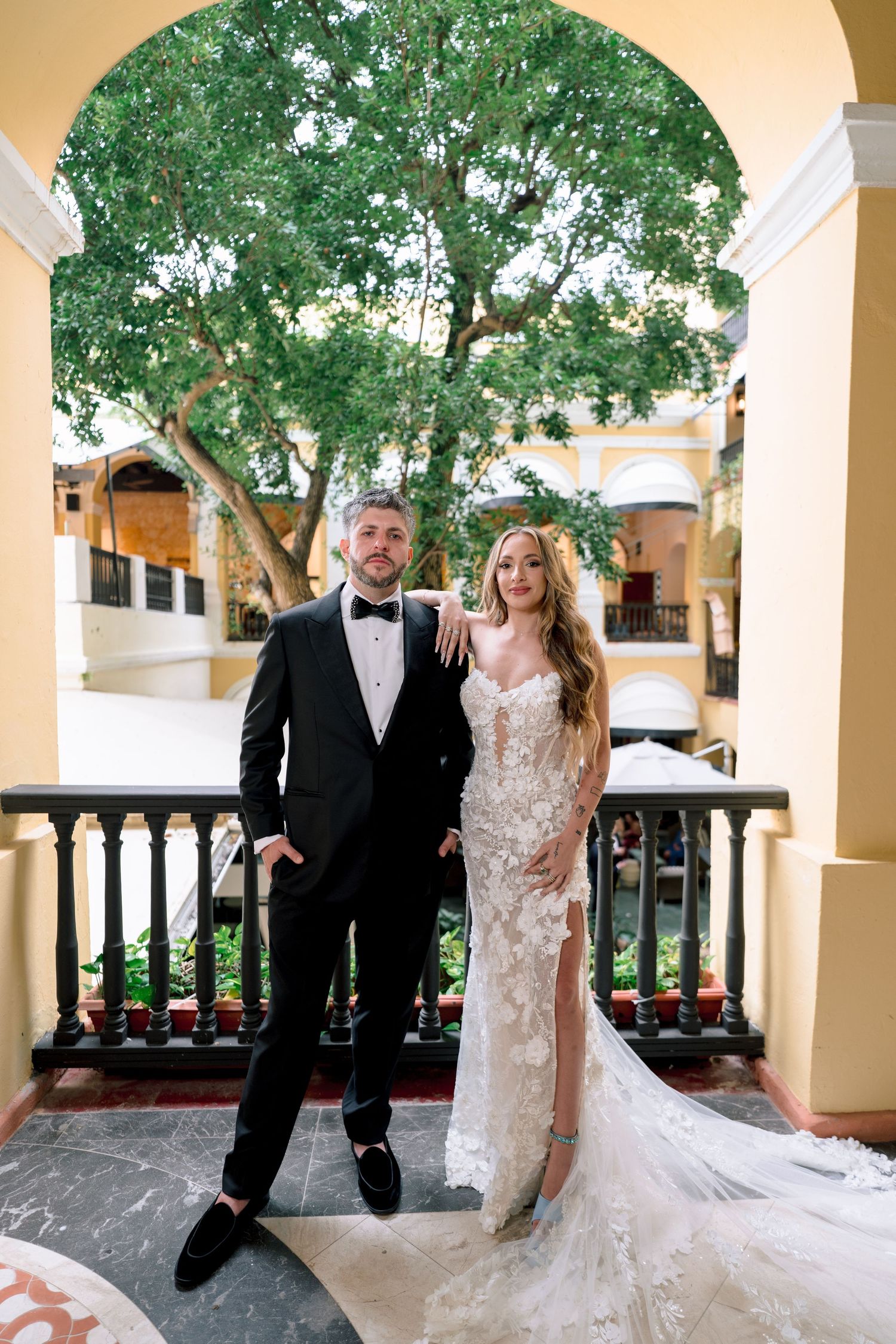 Elegant couple in wedding attire pose on balcony beneath lush green tree arch.