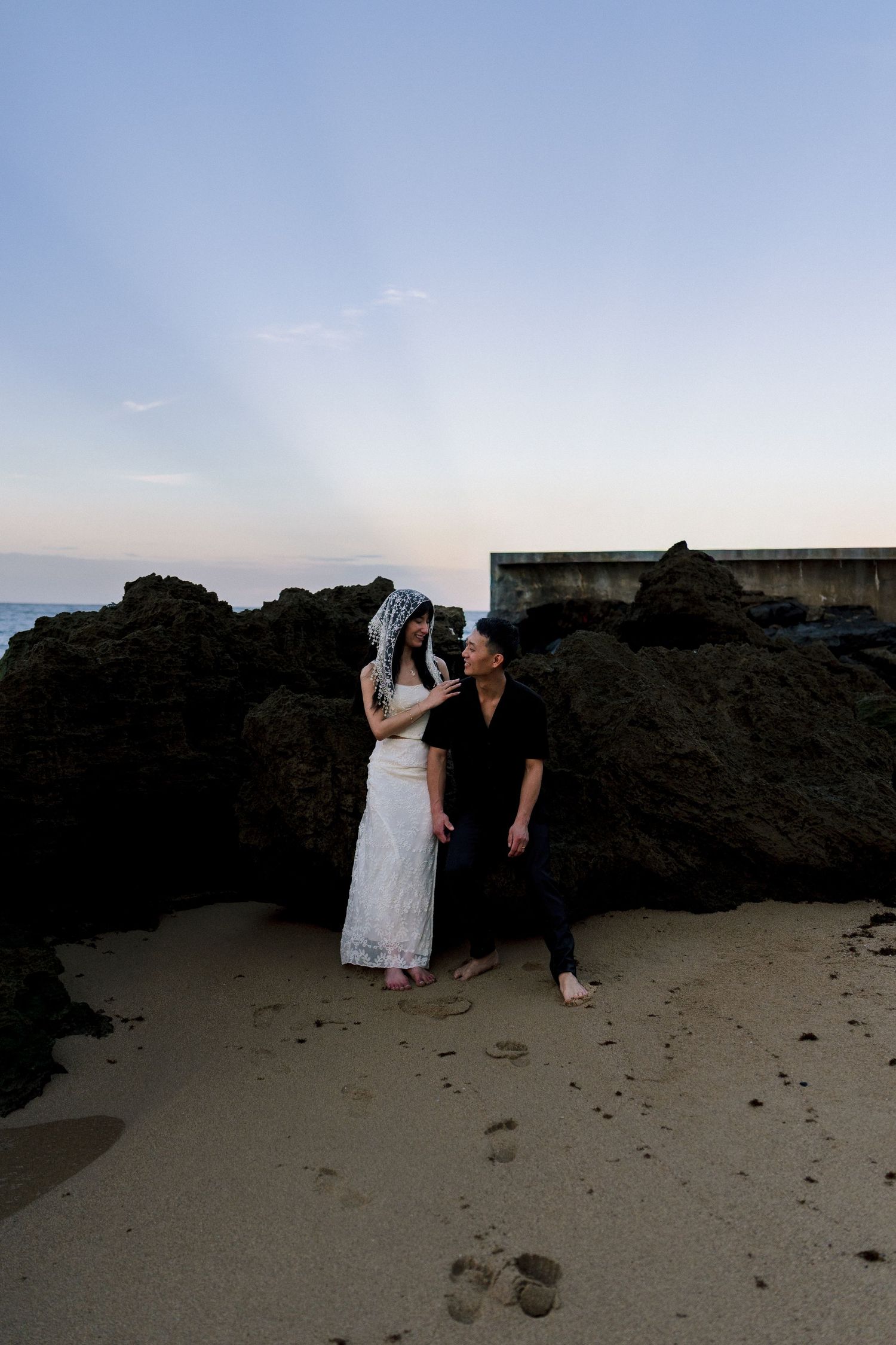 Couple in wedding attire posing on sandy beach at dusk with rocky backdrop and soft twilight sky.