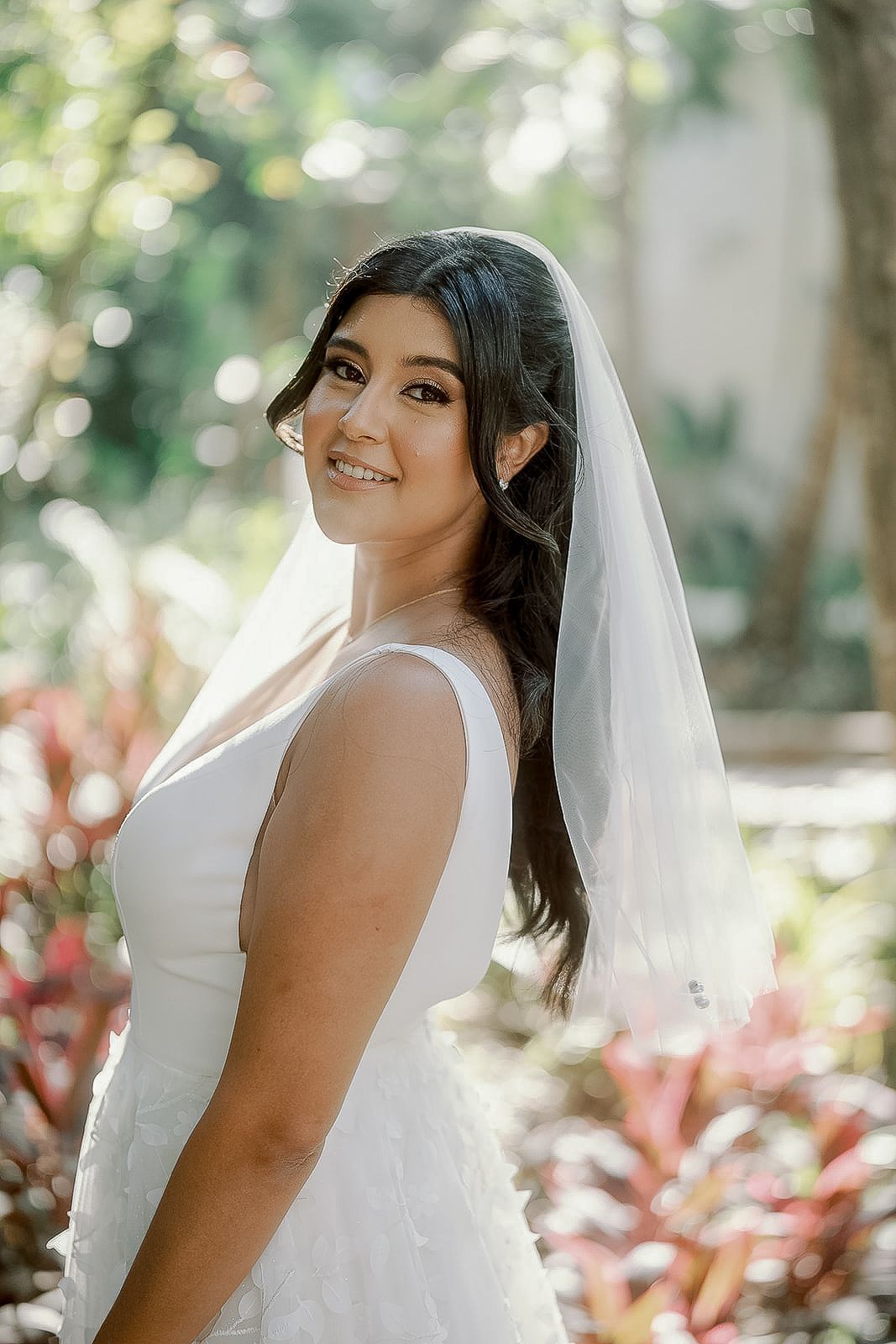 Smiling bride in a white wedding dress and veil posing outdoors in a sunlit garden setting.