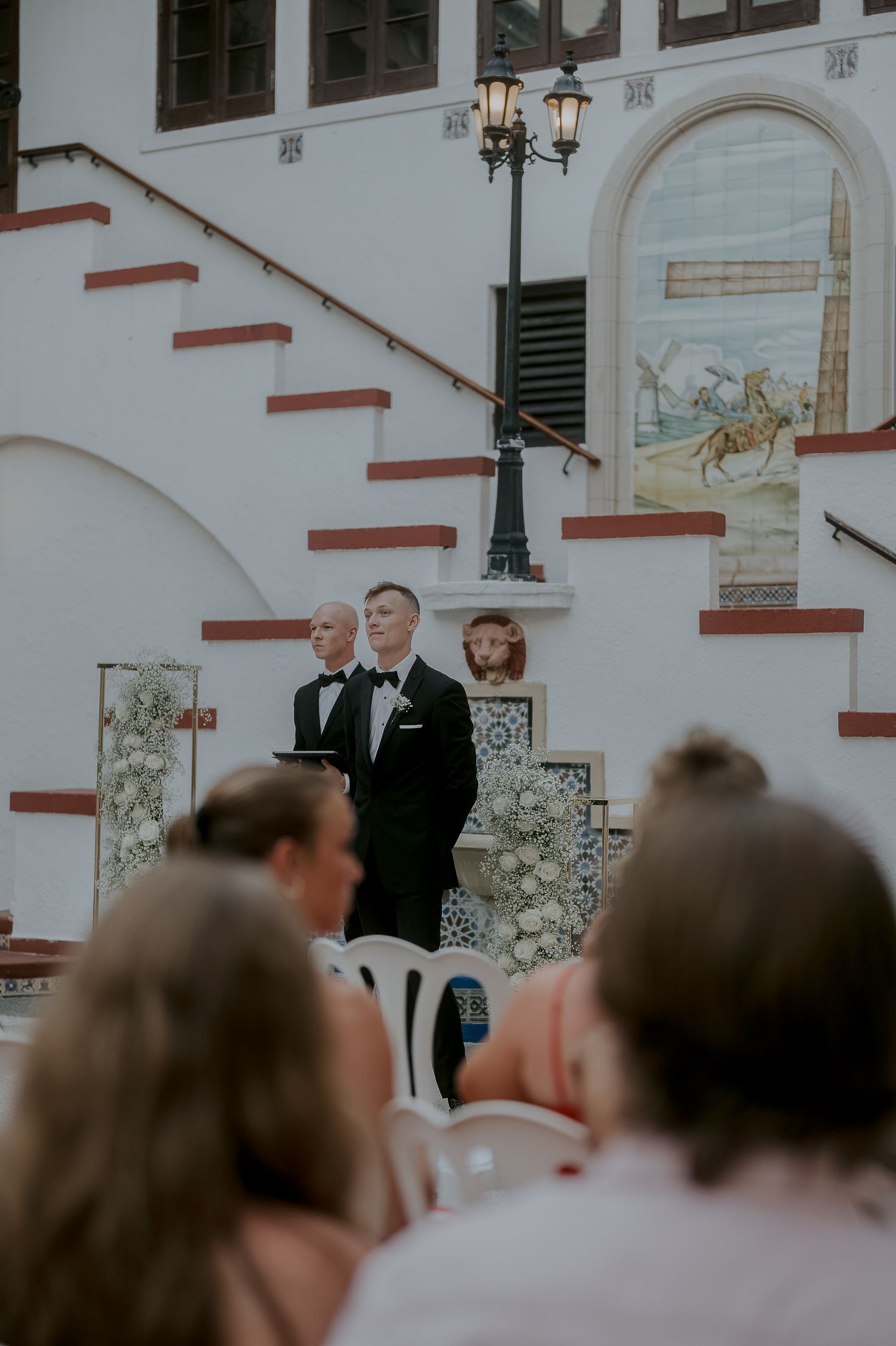 Two grooms in tuxedos stand at outdoor wedding ceremony before ornate tiled building with staircase.