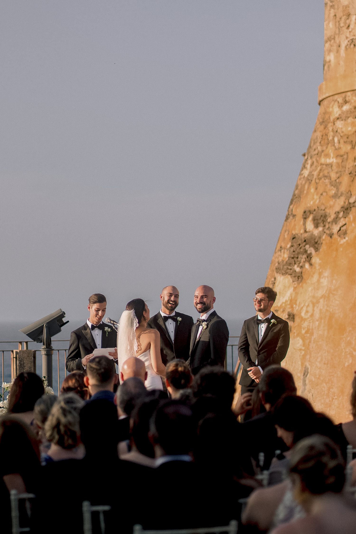 Outdoor wedding ceremony at a coastal fortress with bride, groom, and officiant against a scenic ocean backdrop.