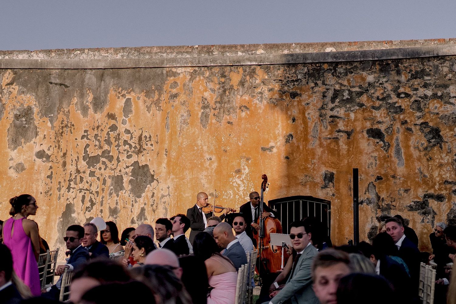 Outdoor wedding reception with live band performing against a weathered yellow wall, guests mingling in elegant attire.
