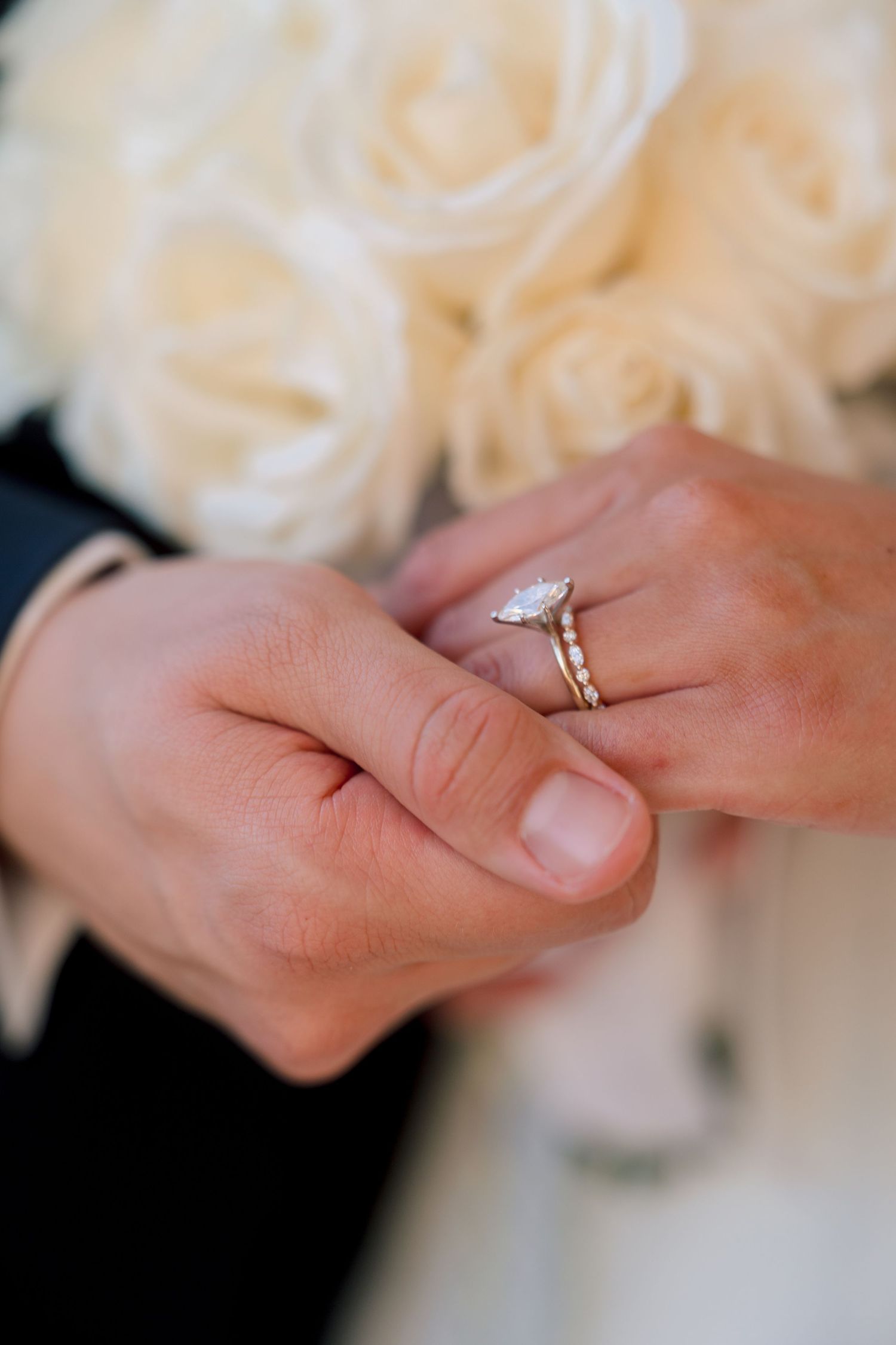 Close-up of bride's hand wearing marquise diamond engagement ring and wedding band, held by groom near white rose bouquet.