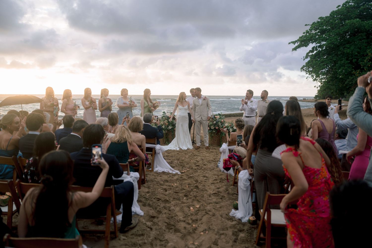 Beach wedding ceremony at sunset with bride and groom exchanging vows, guests seated on sand, ocean backdrop.