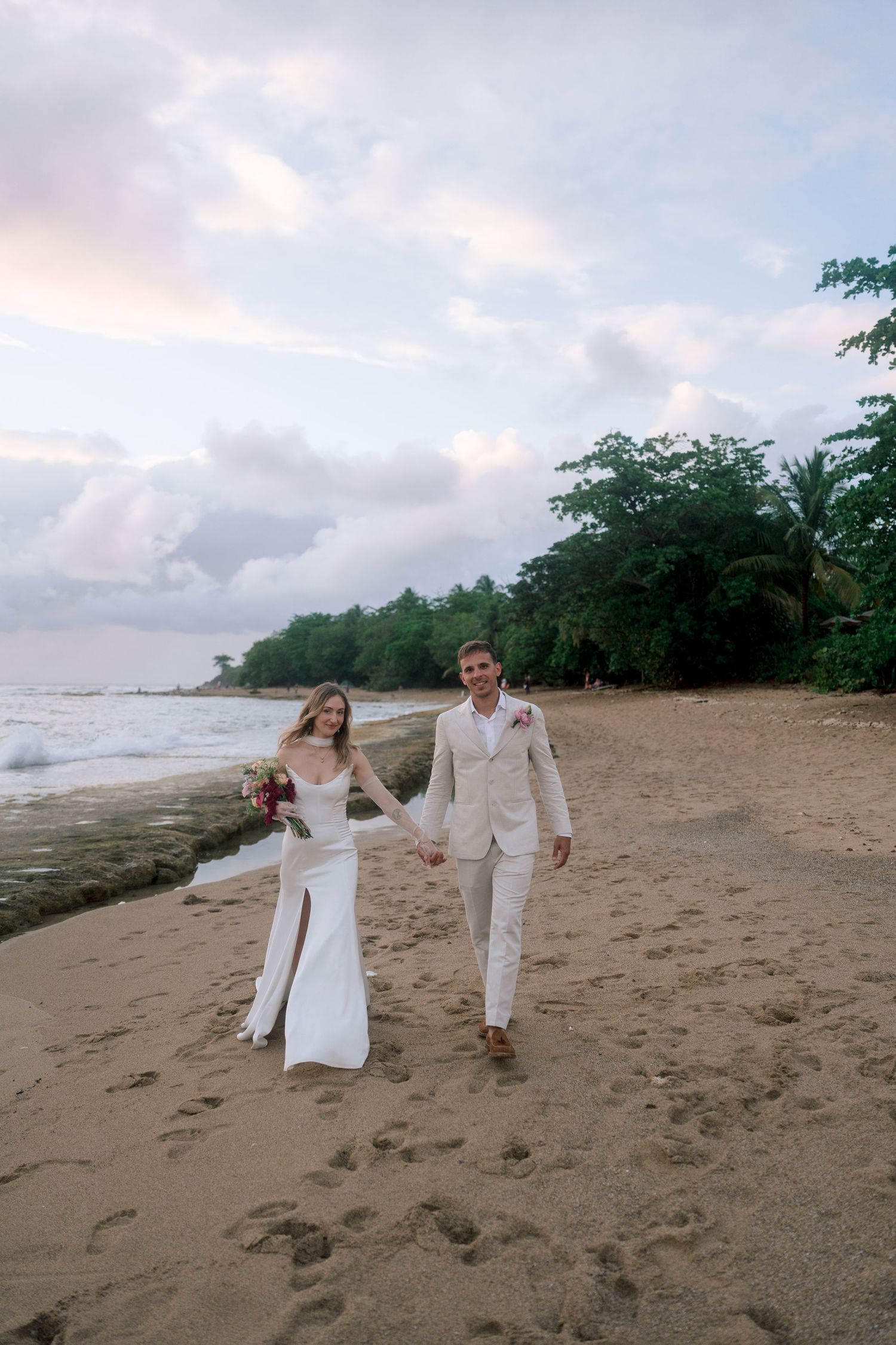 Smiling bride and groom holding hands walking along tropical beach at sunset after wedding ceremony.