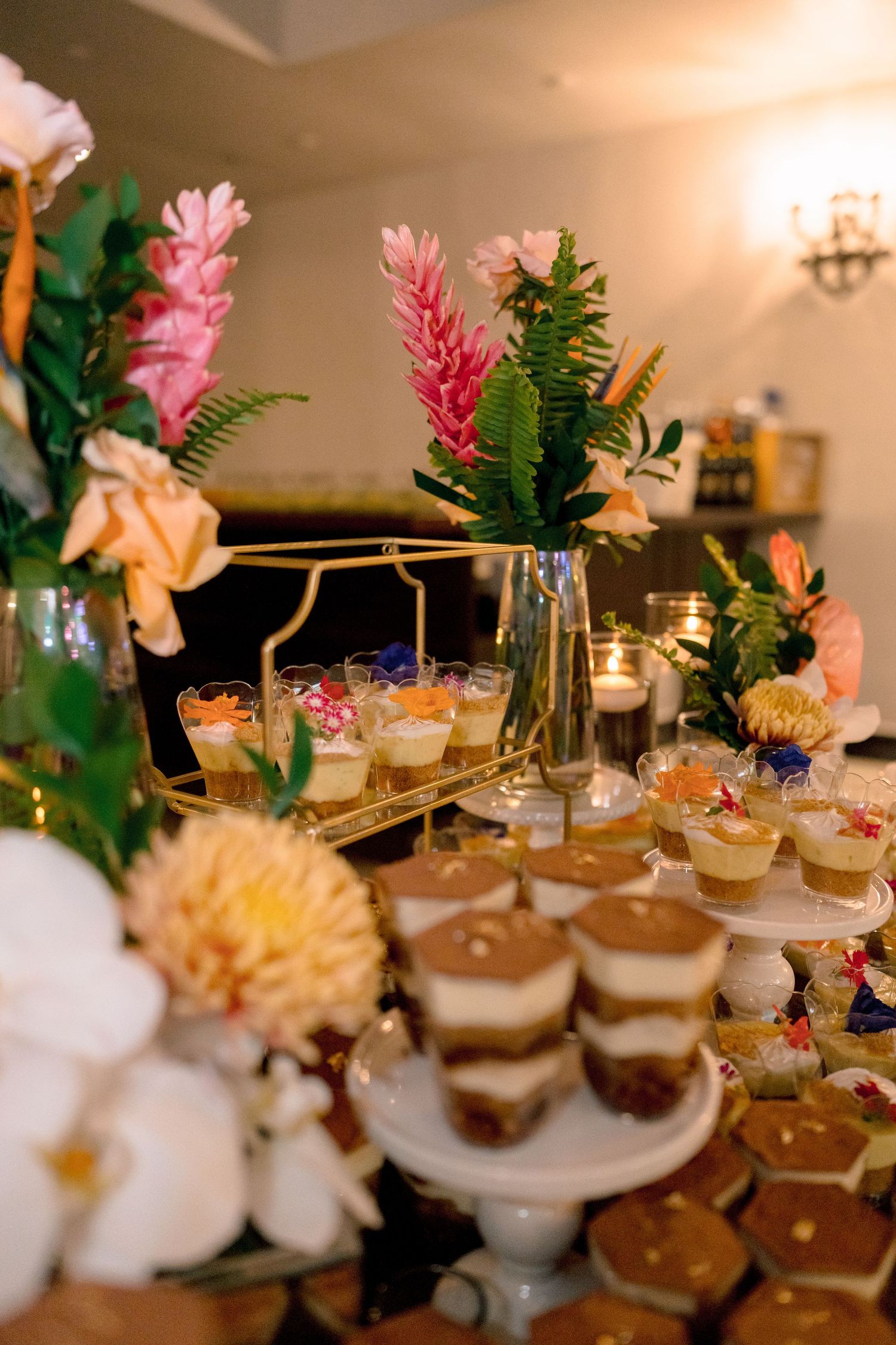 Elegant wedding dessert table with layered tiramisu cups, tropical floral arrangements, and candlelit petit fours.