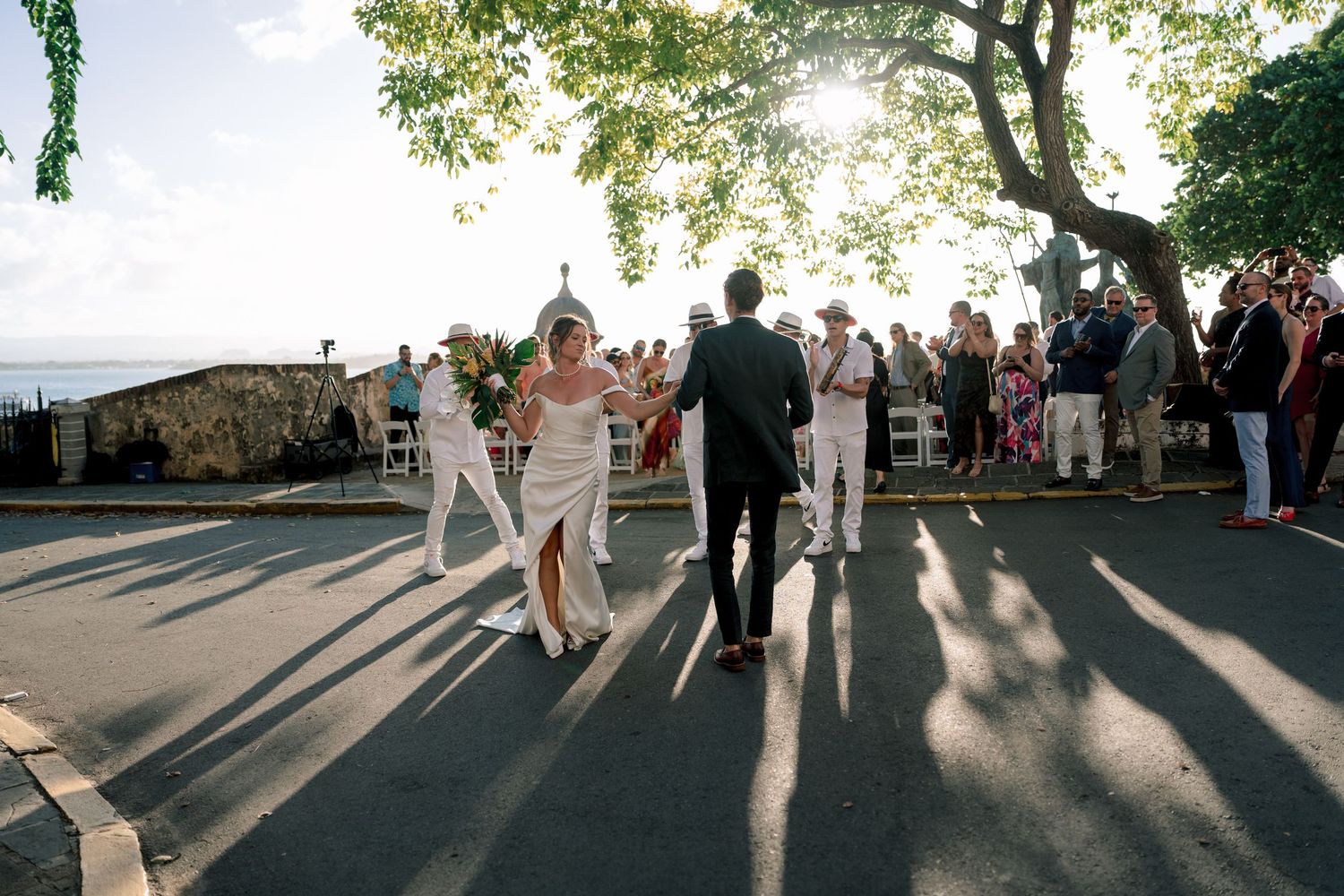 Bride and groom walk together at outdoor waterfront wedding ceremony with guests and tree backdrop.
