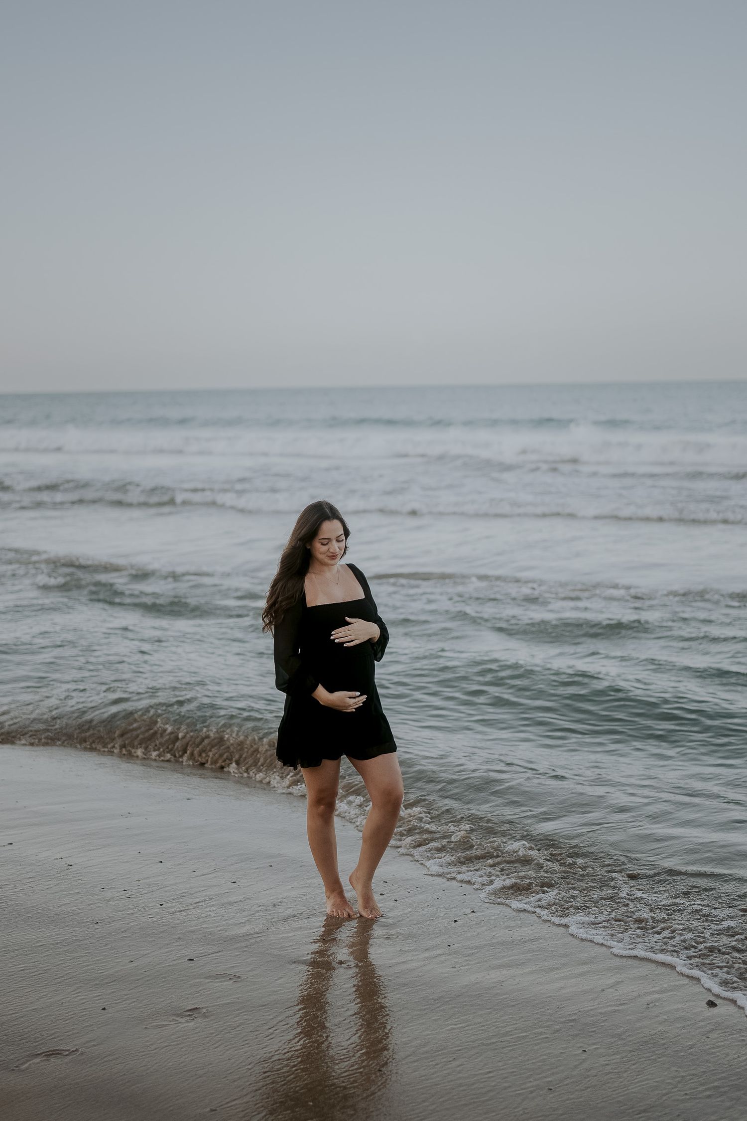 Pregnant woman in black off-shoulder dress cradling baby bump while walking barefoot on a sandy beach.