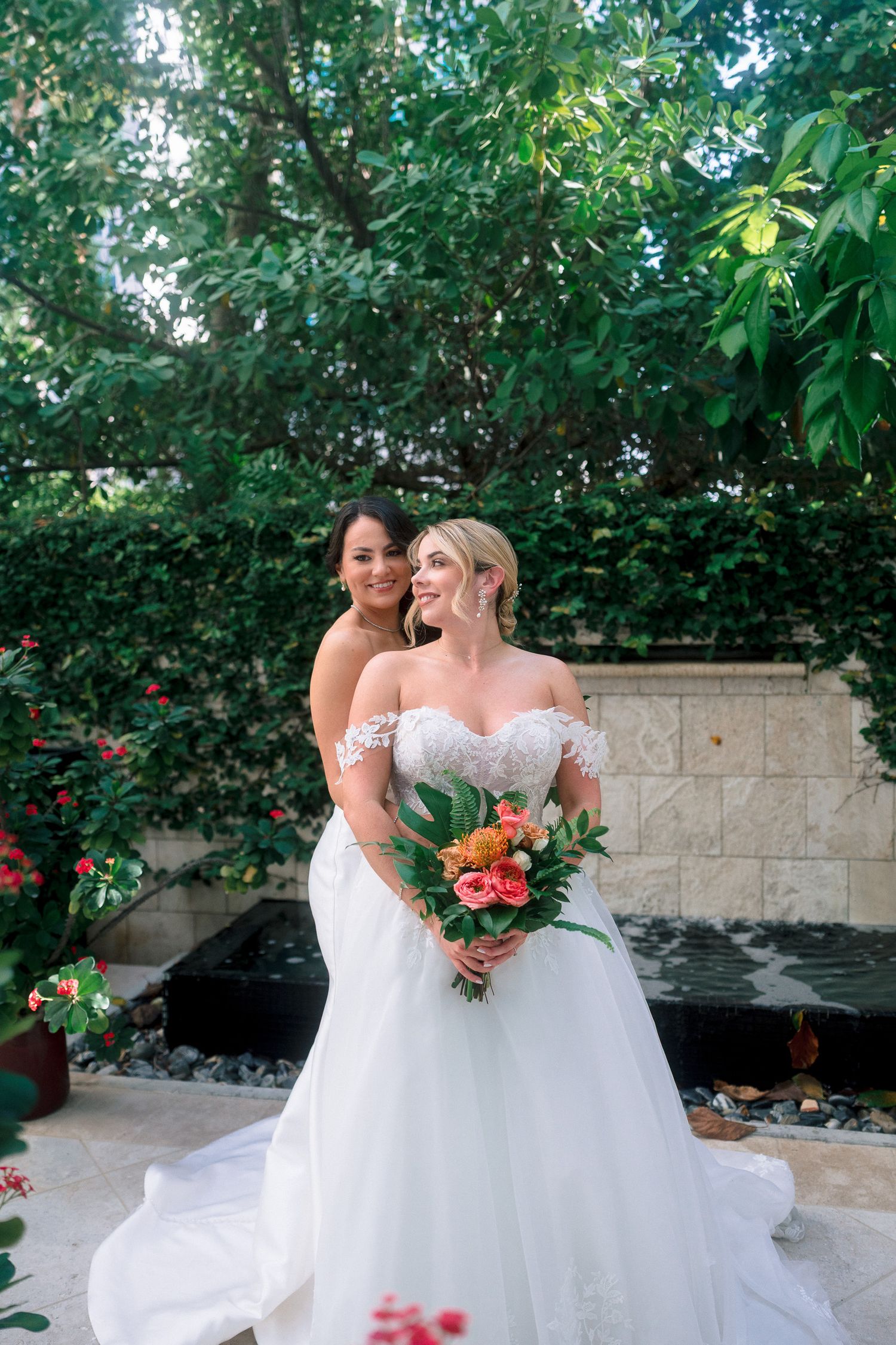Two brides in white wedding gowns pose together outdoors, one holding a vibrant tropical bouquet, surrounded by lush greenery.
